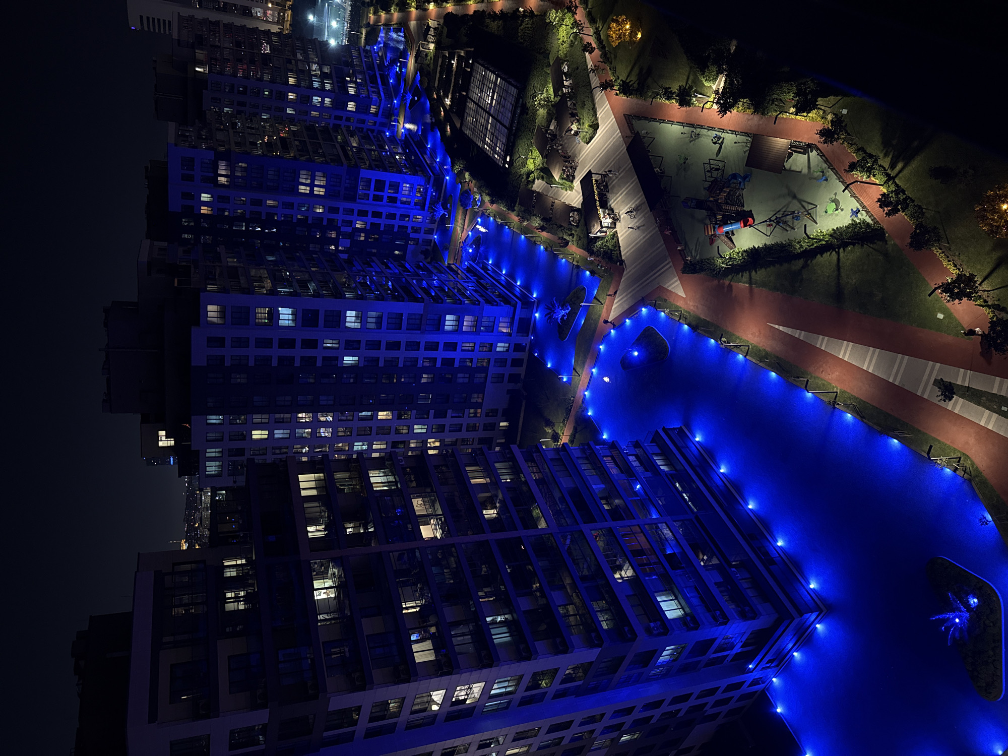 This is a high-angle, full-shot, night-time, outdoor, aerial photograph of a modern apartment complex with blue LED lighting illuminating the lower levels and pathways. The buildings are tall, with many windows showing interior lights, suggesting occupancy. In the foreground, a playground with various colorful equipment is visible, surrounded by green lawn and trees. Pathways, some with a red track surface, wind through the complex. Several small, covered seating areas are scattered throughout the landscaped grounds. A few individuals and bicycles are present in the scene, indicating activity in the common areas. The city of Ümraniye, Turkey, is implied by the architectural style and the context provided. The overall impression is one of a well-lit, modern, and active residential community.