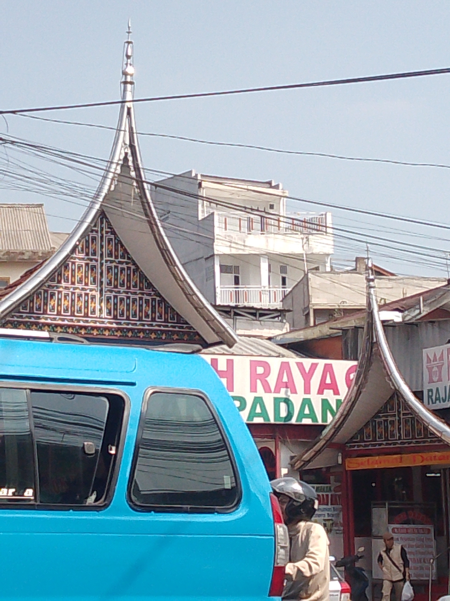 A street scene in Cisarua, Indonesia, features traditional Minangkabau architecture with distinctive curved roofs adorned with intricate geometric patterns. A bright blue van is partially visible in the foreground on the left. Behind the van, a banner with the words "H RAYA PADANG" is displayed. Further back, a multi-story building with balconies stands against a clear blue sky. Overhead, numerous electrical wires crisscross the frame. Two individuals are visible near a shop entrance to the right, with one wearing a beige jacket and helmet, and another standing further back in a vest. The overall impression is of a bustling urban environment with a blend of traditional and modern elements.