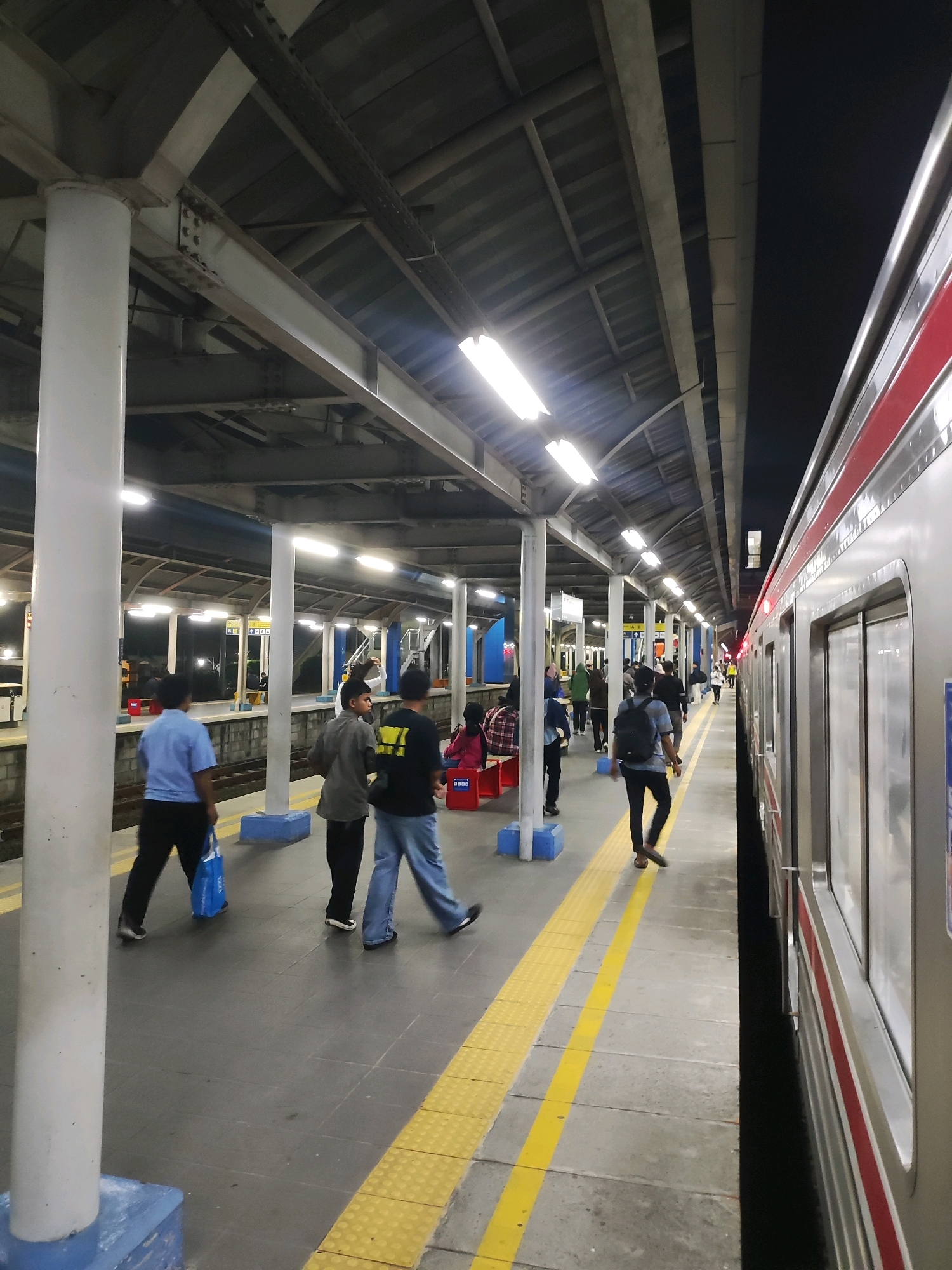 The image captures a nighttime scene at a train station in Tambun, Indonesia. A train is parked alongside the platform, its metallic exterior reflecting the station's artificial lights. Several individuals are present on the platform, walking or standing. One person in a blue shirt and black pants walks away from the camera, carrying a blue bag. Another person in a gray shirt and black pants is walking towards the right side of the frame. A third person, also in a gray shirt, is seen from the back with a backpack. The platform is illuminated by overhead fluorescent lights, casting a cool glow. Yellow lines mark the edge of the platform, and a safety barrier is visible. The station's architecture includes a covered roof supported by white pillars and metallic beams. The background shows the dark night sky and the distant lights of the city.