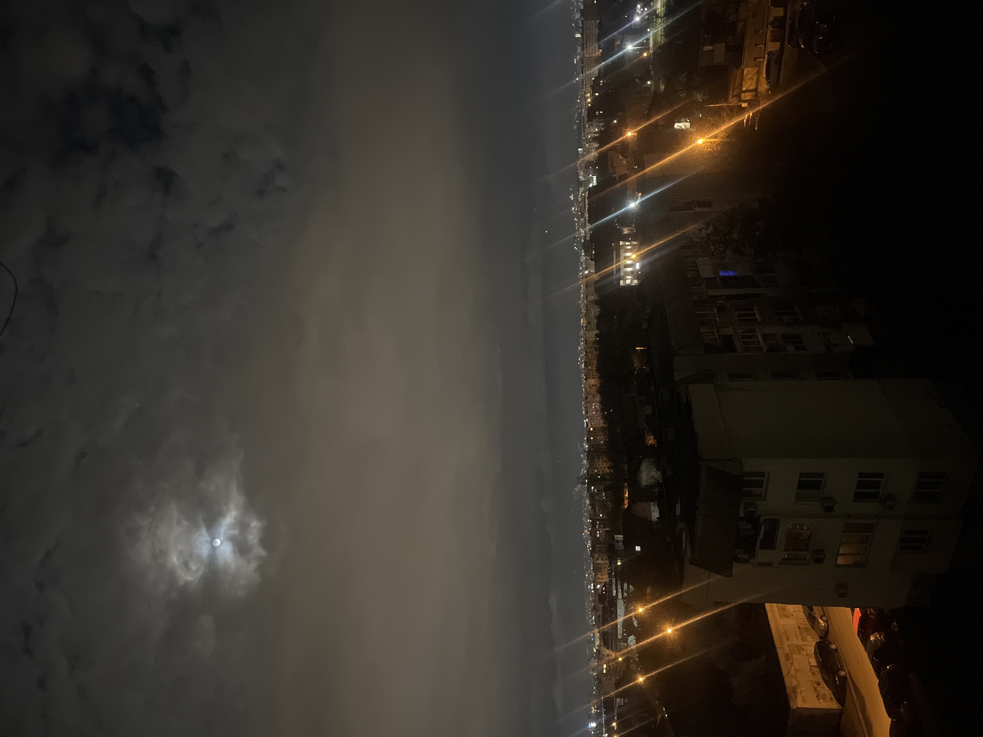 The image shows a nighttime view of a city skyline under a cloudy sky with the moon visible. There are building lights and street lights visible, suggesting an urban setting. Based on the street layout and building styles, as well as the presence of Bulgarian language information in the metadata, the location is likely the city of Sofia, Bulgaria. There are no people visible. The image depicts a nighttime scene with urban infrastructure and atmospheric conditions.