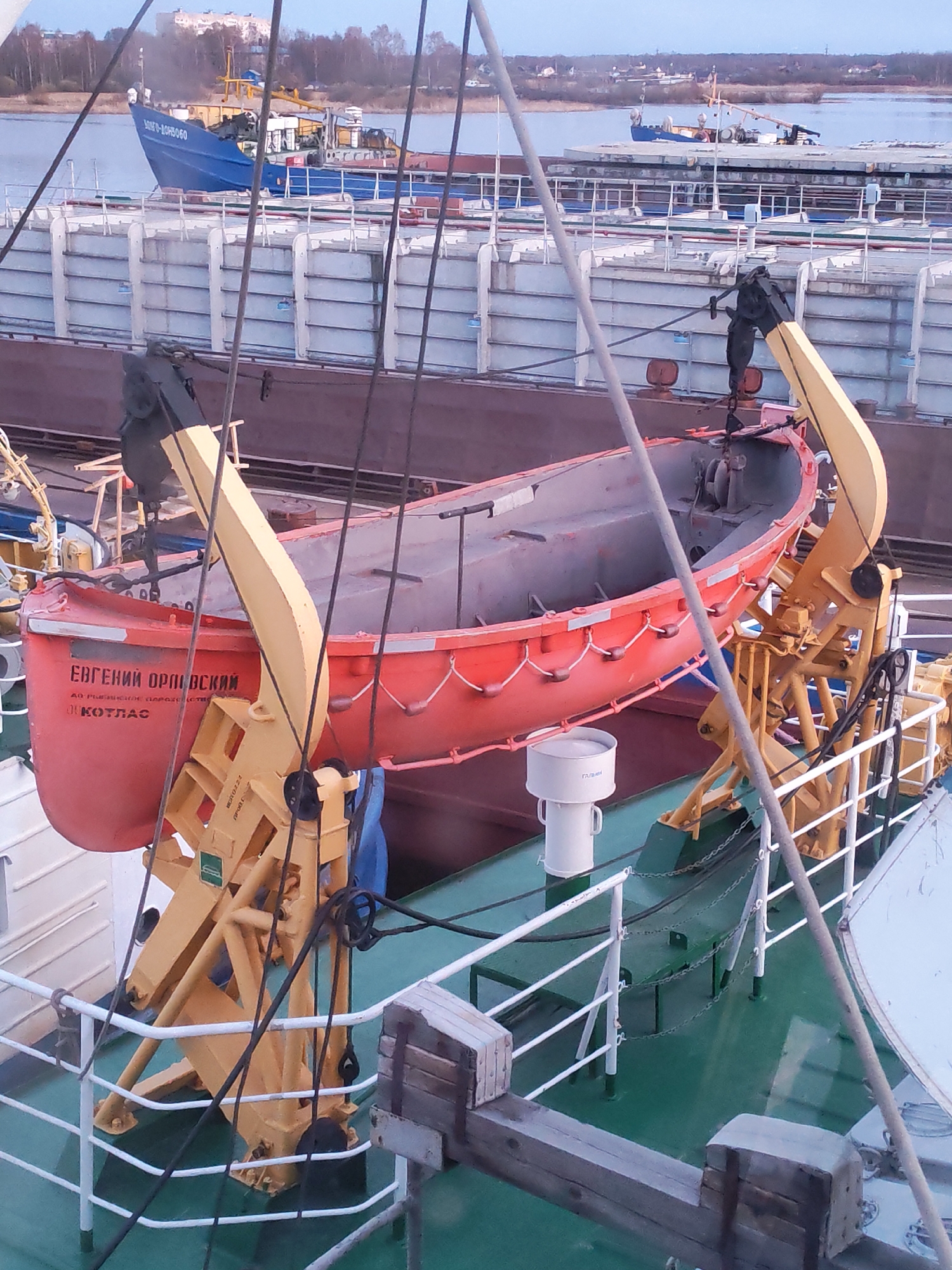 The image shows a red lifeboat labeled "Евгений Орловский" (Evgeniy Orlovskiy) and "КОТЛАС" (Kotlas) held by yellow davits on the deck of a ship. The deck is painted green and white. In the background, another ship, painted blue and labeled "МЗТО-1045060," is visible. The setting appears to be a river or port area. The city of Rybinsk, Russia is not identifiable in this image.