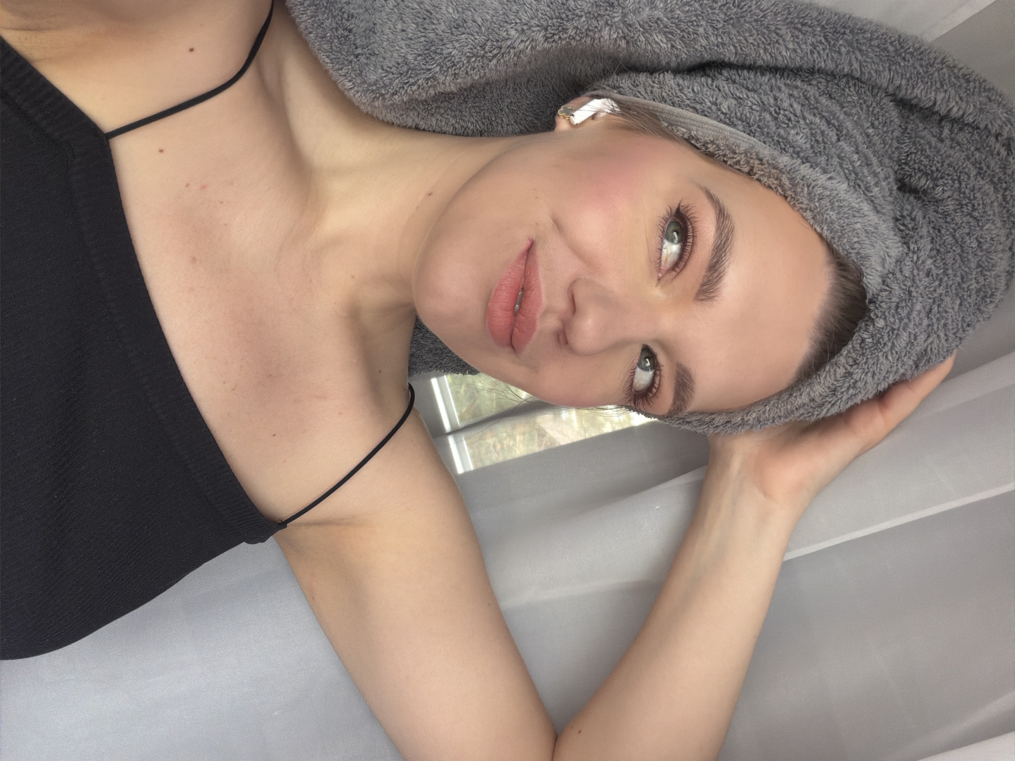 A woman is lying down, facing the camera. She has fair skin, light-colored eyes, and pink lips. A gray towel is wrapped around her head, obscuring her hair. She is wearing a black tank top with thin straps. Her right arm is visible, bent, and appears to be holding the towel in place on her head. A window is visible in the background, but the view outside is obscured by glare. The background is primarily a smooth, gray surface. The location in the city of Dreiliņi, Latvia, cannot be confirmed from the image.