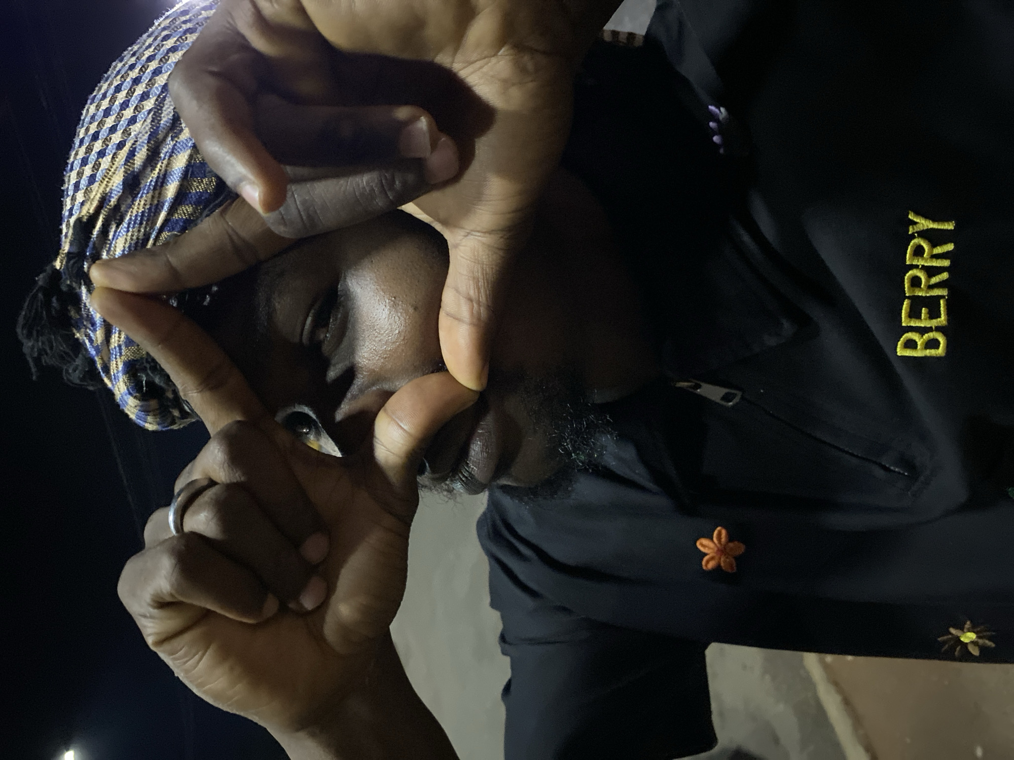 A close-up, waist-up shot shows a person wearing a black shirt with embroidered flowers and the name "BERRY" in yellow letters. The person has dark skin and is wearing a headscarf with a blue, yellow, and white checkered pattern. Their hands are positioned in front of their face, with fingers forming a frame around their eyes. The background is dark, suggesting it is nighttime, and a street light is visible in the upper left corner. This image was captured outdoors at night in Bukuru, Nigeria.