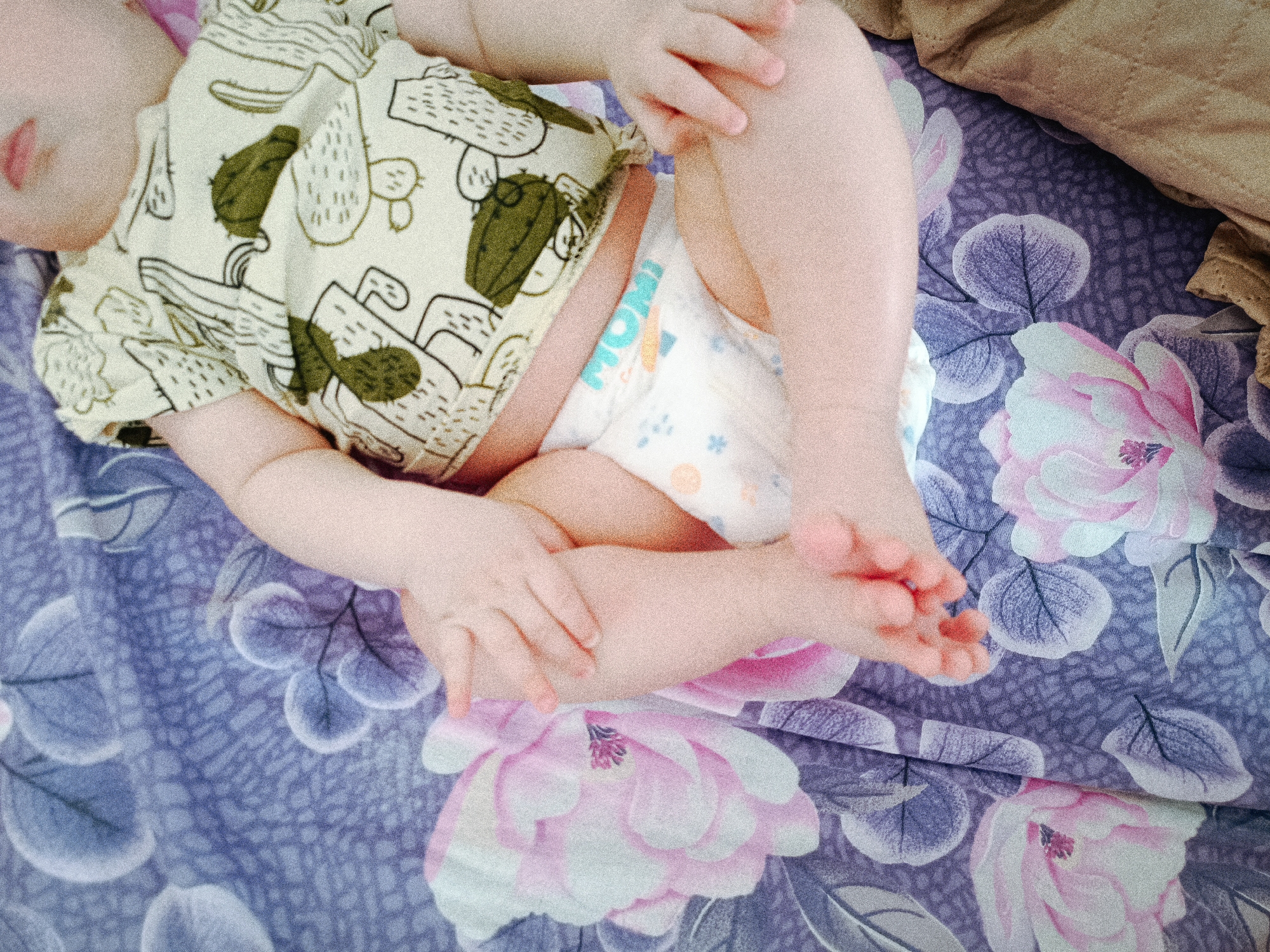 A baby lies on a bed covered with a floral-patterned sheet. The baby wears a diaper and a cactus-themed shirt. The location is not discernable from the image provided, and therefore the city of Tobolsk, Russia, cannot be confirmed.