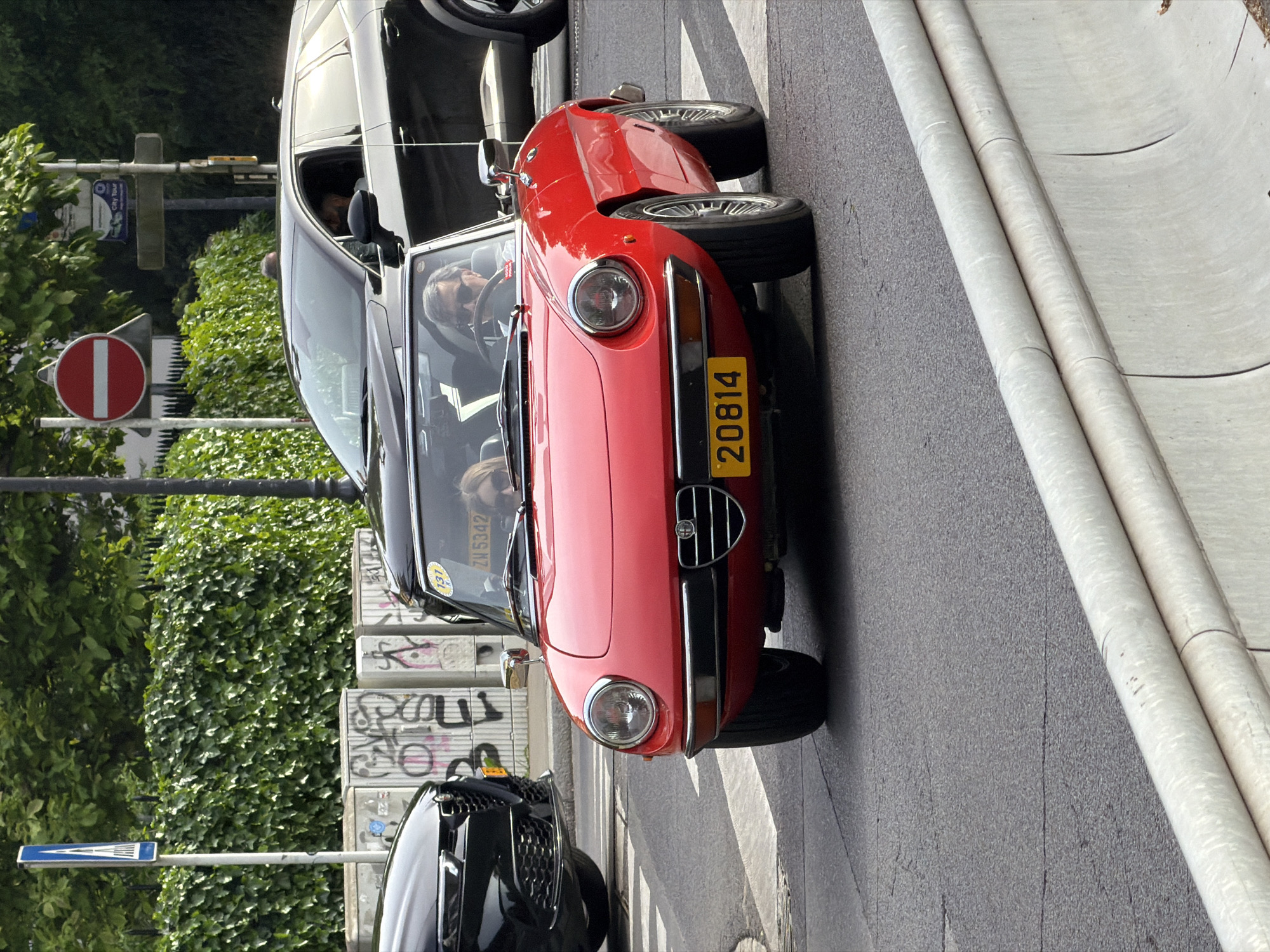 A ground-level medium shot shows a red Alfa Romeo Spider car with license plate "20814" stopped in a crosswalk in the city of Luxembourg, Luxembourg. The license plate on the windshield is “ZN5342”, and a sticker on the windshield reads “131”. The driver is a man in his fifties or sixties with gray hair, looking forward. The passenger is a woman in her twenties or thirties. To the car's left is a black vehicle stopped at the crosswalk. To the left of the vehicles are a graffiti-covered wall, green foliage, and a street sign. A "no entry" sign is also visible. To the right is a curb. The shot is slightly tilted. The weather appears to be sunny.
