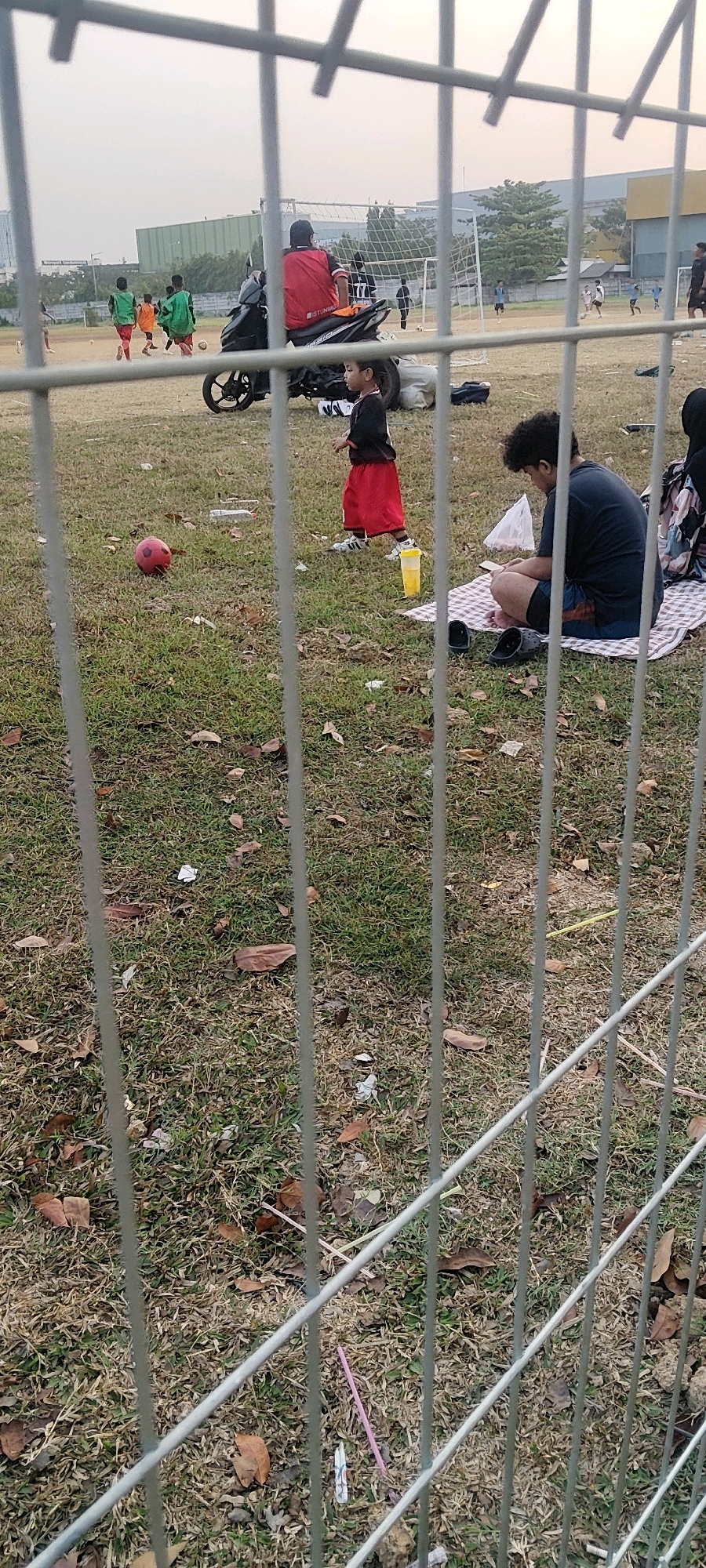 The image depicts an outdoor scene at a grassy field, likely a sports field given the presence of a soccer ball and goalposts in the background. The foreground is dominated by a metal fence.
In the middle ground, a young boy wearing a black shirt with red trim and red shorts is standing and looking to his left. A red soccer ball is on the grass to the left of the boy. Further back, several young boys in green shirts are playing soccer, with a person in a red vest on a motorcycle observing them. In the background, more individuals are visible on a soccer field, and buildings can be seen beyond the field.
To the right of the young boy, a person is sitting on a checkered blanket on the grass, looking down at something in their lap. There are shoes on the ground near them, and a yellow cup is also present. The ground is covered in dry grass and fallen leaves, suggesting a dry season.
The sky is overcast and pale. The location is identified as Pasirsari, Indonesia.