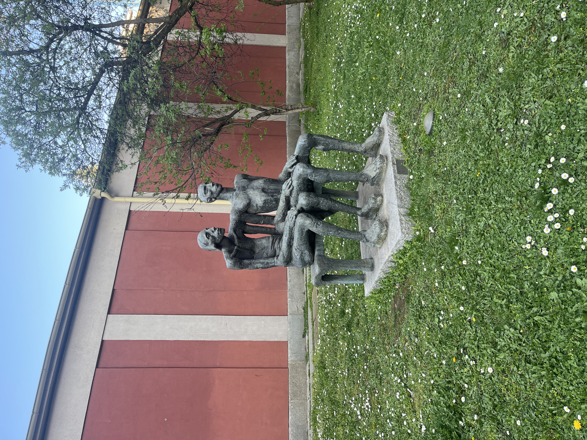 The image depicts a bronze sculpture of three seated figures located outdoors, with a building and grass in the background. The sculpture, likely depicting human figures, has a weathered appearance with a green patina. The figures are huddled together, with discernible facial features.
The building in the background has a red facade with a light gray horizontal stripe. A tree is visible behind the sculpture.
The foreground is filled with green grass, interspersed with small white flowers.
Based on visual cues and context, the sculpture is likely located in a public space in the city of Sofia, Bulgaria.