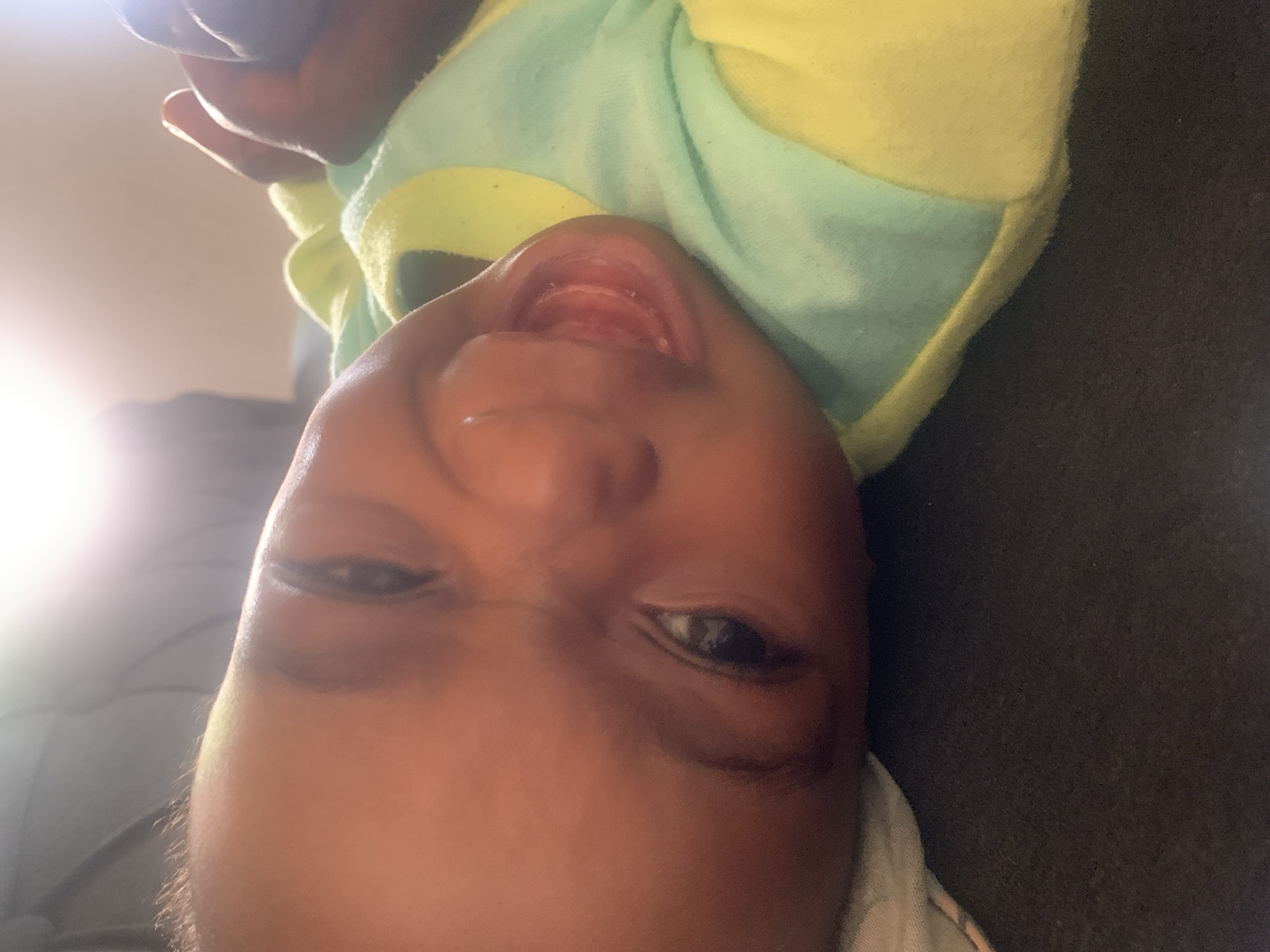 A close-up, upside-down image shows a baby with brown skin wearing a yellow and mint green shirt. The baby's mouth is open, revealing the upper gums. The background is partially blurred, showing a dark gray surface on one side and a lighter gray or white surface with sunlight on the other. No information regarding location or action is observable.