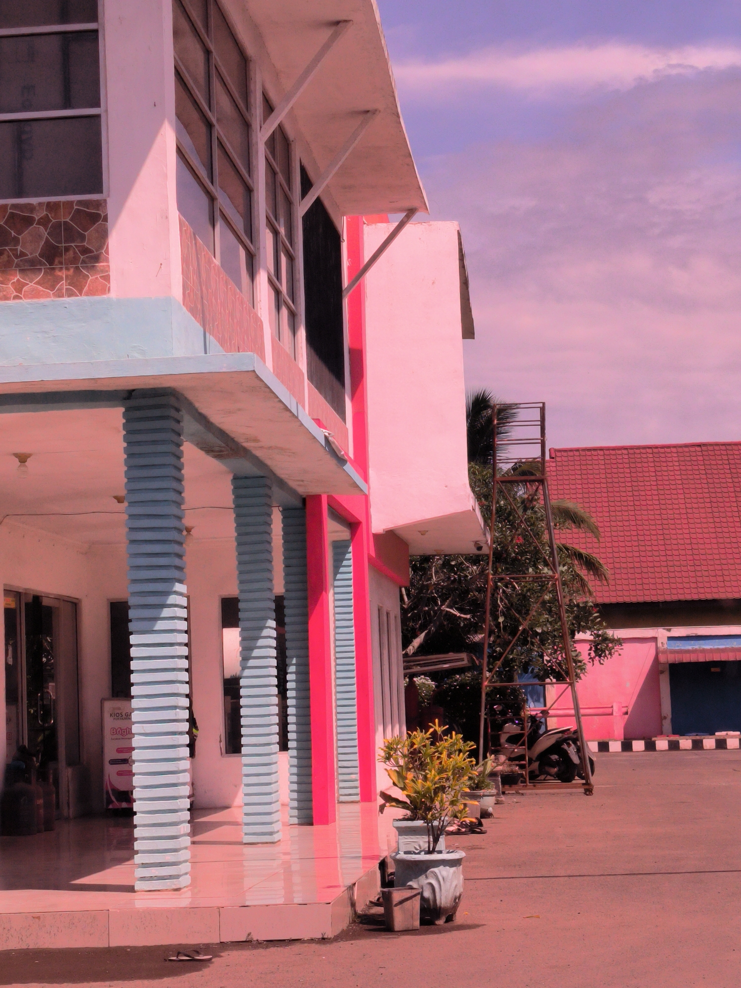 This outdoor photograph depicts a building in Cotpuuk, Indonesia, rendered with a distinctive pinkish-purple color cast affecting the entire scene, including the sky and ground. The prominent structure on the left is a two-story building with a wide overhang supported by several light blue, horizontally ribbed columns and a few solid red columns. The upper floor features multiple large windows with white frames, and the lower sections of the building are adorned with brown and reddish mosaic-like tiling, as well as a light blue painted base.
In the mid-ground, a paved area extends forward from the building, showing reflections on its shiny surface. A large potted plant with yellow-green foliage stands prominently on the right side of the foreground, with other smaller potted plants receding into the distance along the building's facade. A single flip-flop is visible on the ground near the prominent potted plant.
Further back on the right, a tall, rusty metal structure, resembling a ladder or scaffolding, stands next to the main building. Behind this, a motorcycle (scooter type) is parked. In the background, another building with a striking red tiled roof and reddish-pink walls is visible, featuring a blue door or shutter. Black and white checkered markings line a curb in front of this background building. Lush green trees and palm fronds are scattered throughout the scene, providing contrast to the building colors.
No people are visible in the image. The bright, even lighting and cast shadows suggest it is daytime, possibly morning or afternoon. The sky above is a soft, cloudy purple-pink, consistent with the overall color grading. Inside the left building's entrance, a white sign is partially visible, displaying the text "KIOS GA..." and below it "...Bright...". The scene is static, capturing a moment in an urban or semi-urban environment.