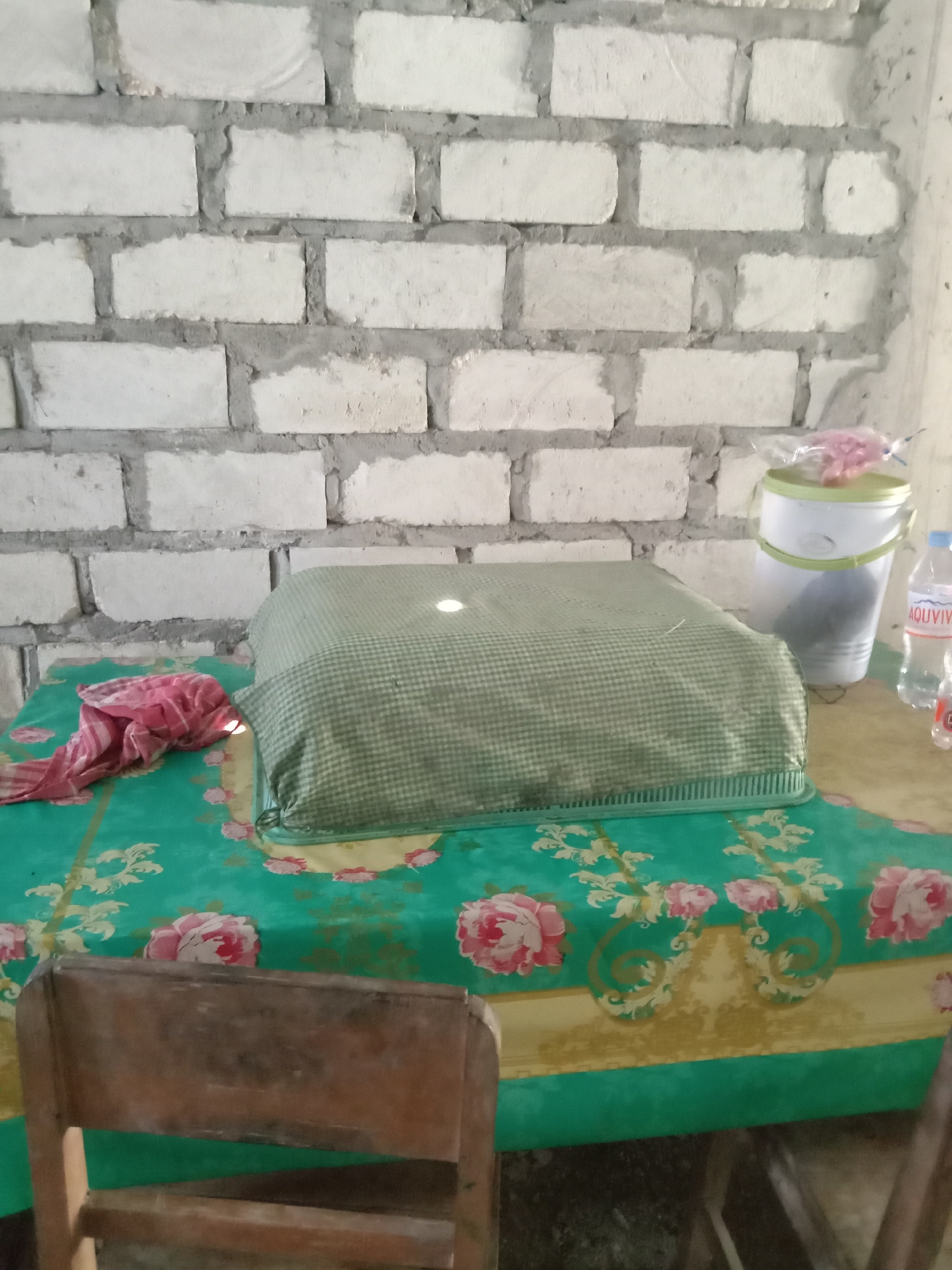 The image depicts an indoor scene with a brick wall as the background. In the foreground, a table is covered with a green tablecloth featuring a floral pattern. On the table, there is a large object covered with a green checkered cloth, and to its left, a crumpled pink fabric. To the right of the covered object, a white bucket with a green lid is visible, and next to it, a plastic bottle of water. A wooden chair is partially visible in the lower left corner. The scene appears to be set in a simple, possibly domestic, environment in Stiaji, Indonesia, judging by the provided location context.