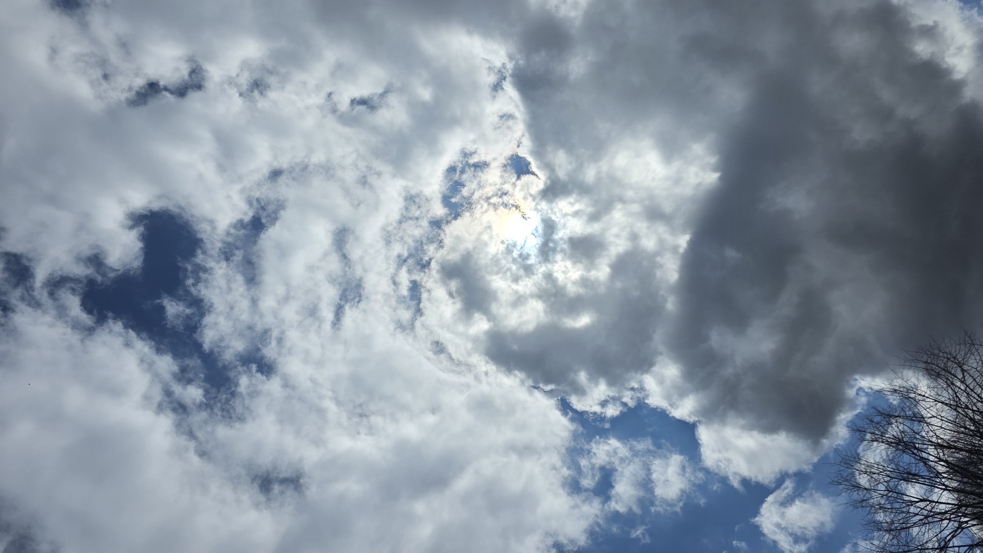 The image shows a sky filled with clouds, with some areas of blue sky visible. The sun is partially obscured by the clouds, creating a bright spot. In the lower right corner, there are bare tree branches. The location in the city of Blagoevgrad, Bulgaria, is not discernible from the image alone.