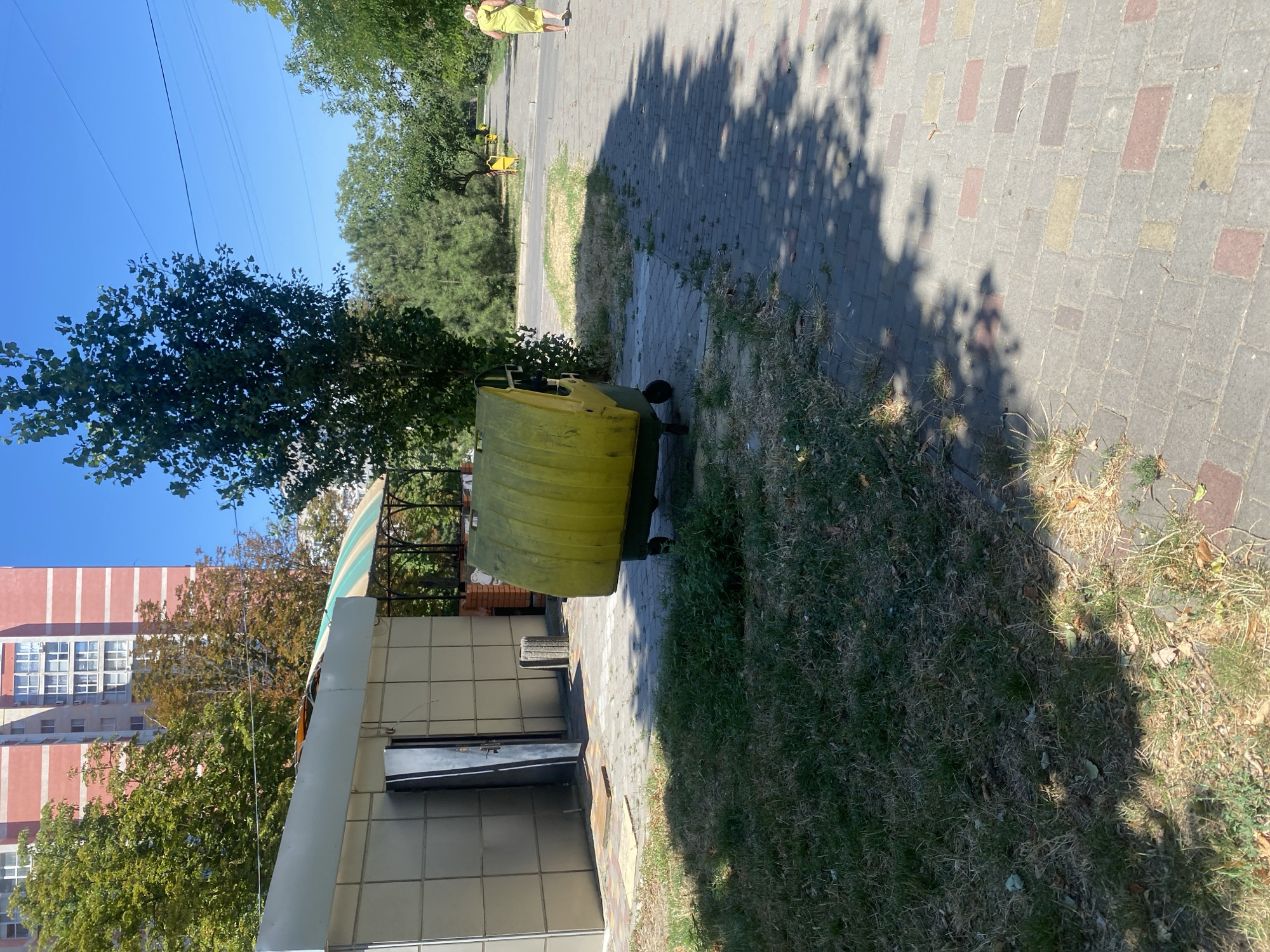 The image shows a yellow dumpster on a paved walkway in Chornomorsk, Ukraine. A woman in a yellow dress walks away from the camera on the walkway, which is lined with trees and grass. In the background, a tall building and a smaller structure with a striped awning are visible. The sky is clear and blue.