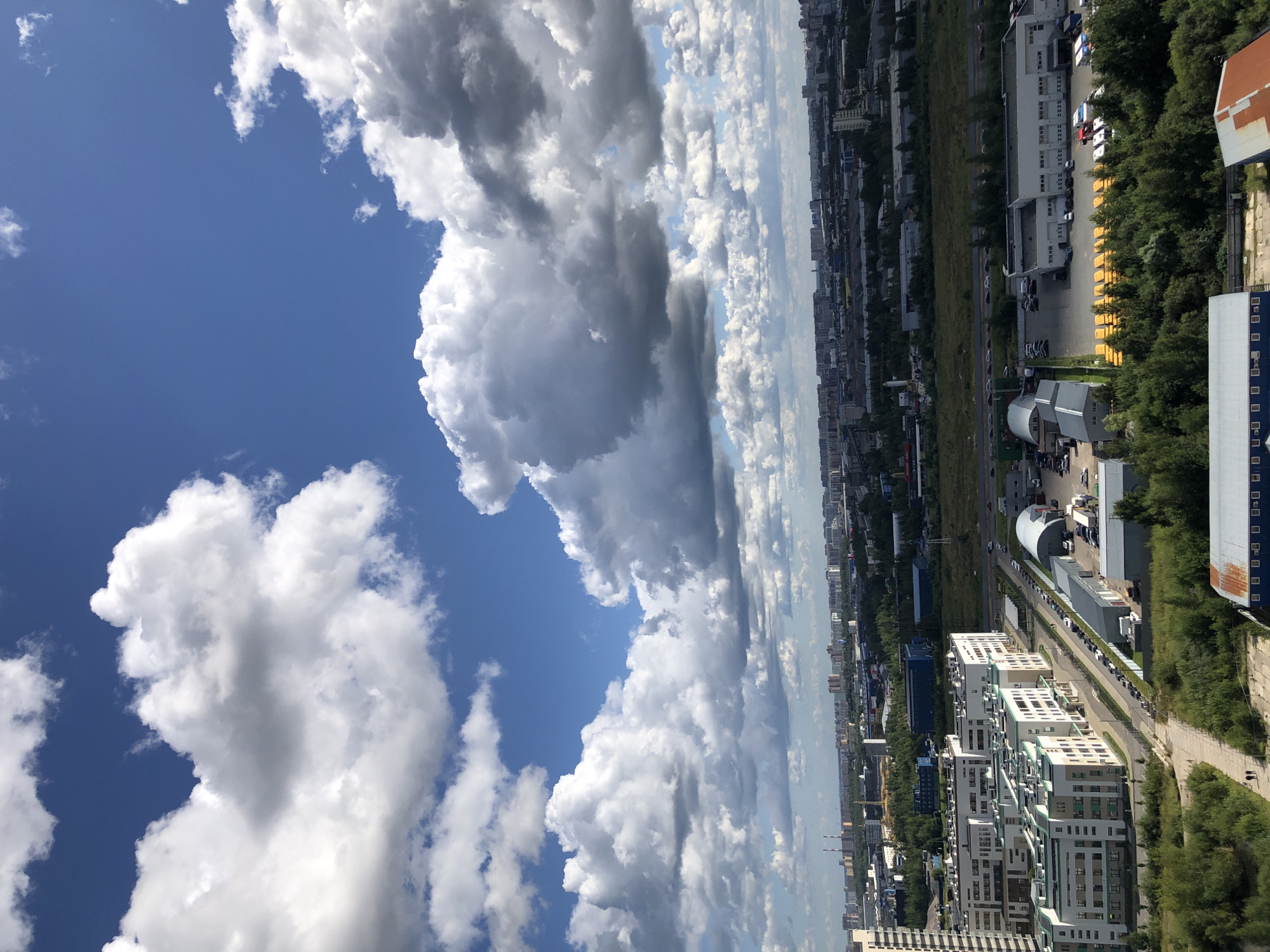 This is an aerial shot of a city skyline on a partly cloudy day. The sky is a vibrant blue with numerous large, fluffy white clouds. Below the sky, a sprawling urban landscape stretches to the horizon, characterized by a mix of residential buildings, industrial complexes, and green spaces. The foreground features modern multi-story apartment buildings with light-colored facades and green accents. A road runs alongside these buildings, with parked cars and trucks visible. Further into the scene, a collection of yellow school buses is parked in front of a long, white industrial building. The background shows a dense urban area with taller buildings and industrial structures, interspersed with trees. The overall impression is of a developed city under a dynamic sky.