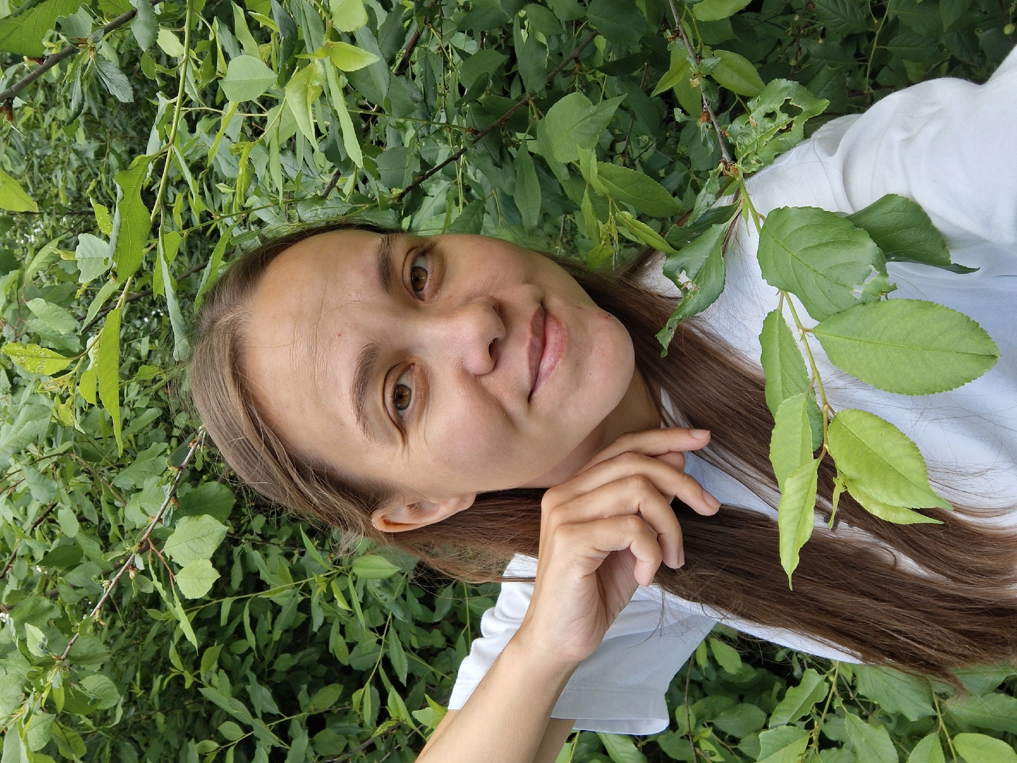 A young adult woman, likely in her 20s or 30s, is featured in a close-up, outdoor portrait. She has long, straight brown hair, brown eyes, and fair skin, looking directly at the camera with a gentle, slightly smiling expression. She is wearing a white short-sleeved top or t-shirt. Her left hand is gently placed with fingers resting near her chin or jawline.
The woman is surrounded by a dense backdrop of lush green foliage, suggesting she is amidst bushes or trees in a natural setting like a garden or forest. The leaves appear vibrant and healthy, indicating a spring or summer season, though some show minor signs of insect damage, such as small holes. The lighting is natural daylight, providing a soft and even illumination. The overall mood of the image is calm and serene. There is no visible text in the image. This scene is located in Chaikovsky, Russia.