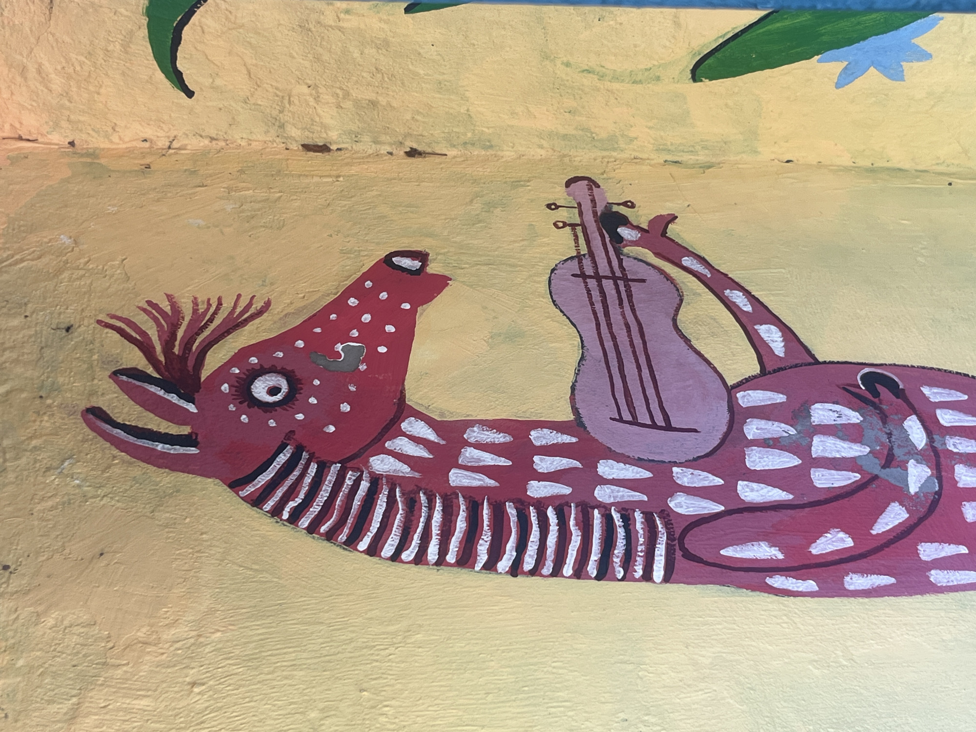 This is a photograph of a whimsical mural painted on a textured yellow wall. The mural depicts a stylized, elongated red creature resembling a horse or a llama, with white polka dots and white, pointed markings along its body and neck. The creature has large, circular eyes with black pupils and white highlights, and a tuft of red hair on its head. It is holding a light purple stringed instrument, similar to a ukulele or a small guitar, with both hands. The instrument has dark horizontal lines representing strings. The background is a textured yellow wall, with a hint of a green leaf and a light blue flower on the right side of the frame. The style of the painting is naive and folk-art inspired. This artwork is located on the wall of a building in Kyiv, Ukraine, likely on a street or public space.
