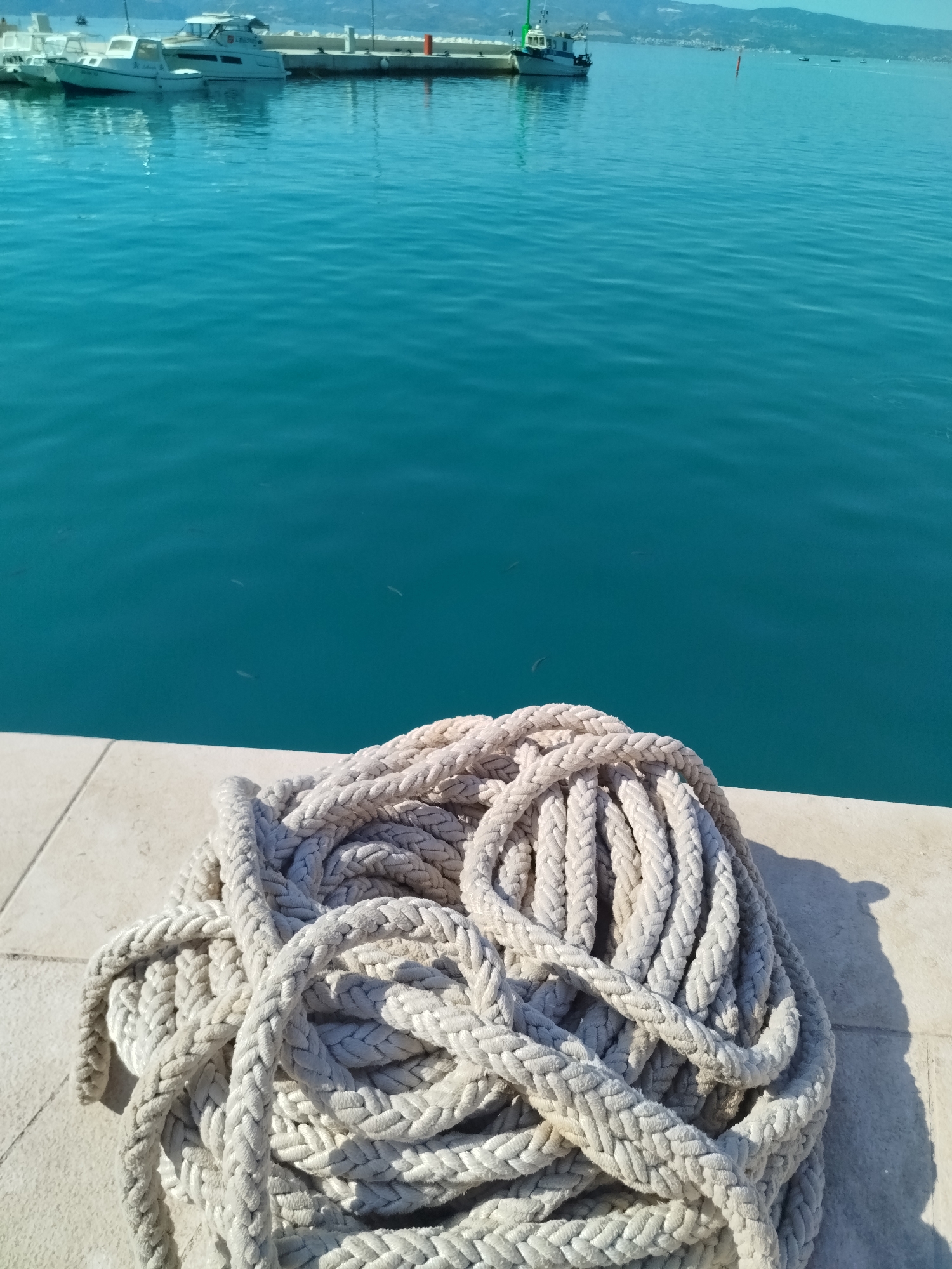 The image depicts a coiled, thick white rope resting on a light-colored tiled surface, likely a pier or waterfront. The rope is substantial and appears weathered. Behind the rope, a vast expanse of bright turquoise water fills the majority of the frame. In the distance, several boats are docked or anchored along a shoreline. The background shows a hazy coastline with hills or mountains under a clear sky. The overall scene suggests a sunny day at a harbor or marina, possibly in the city of Omiš, Croatia, given the characteristic clear blue waters and Mediterranean coastal landscape. There are no people visible in the image.