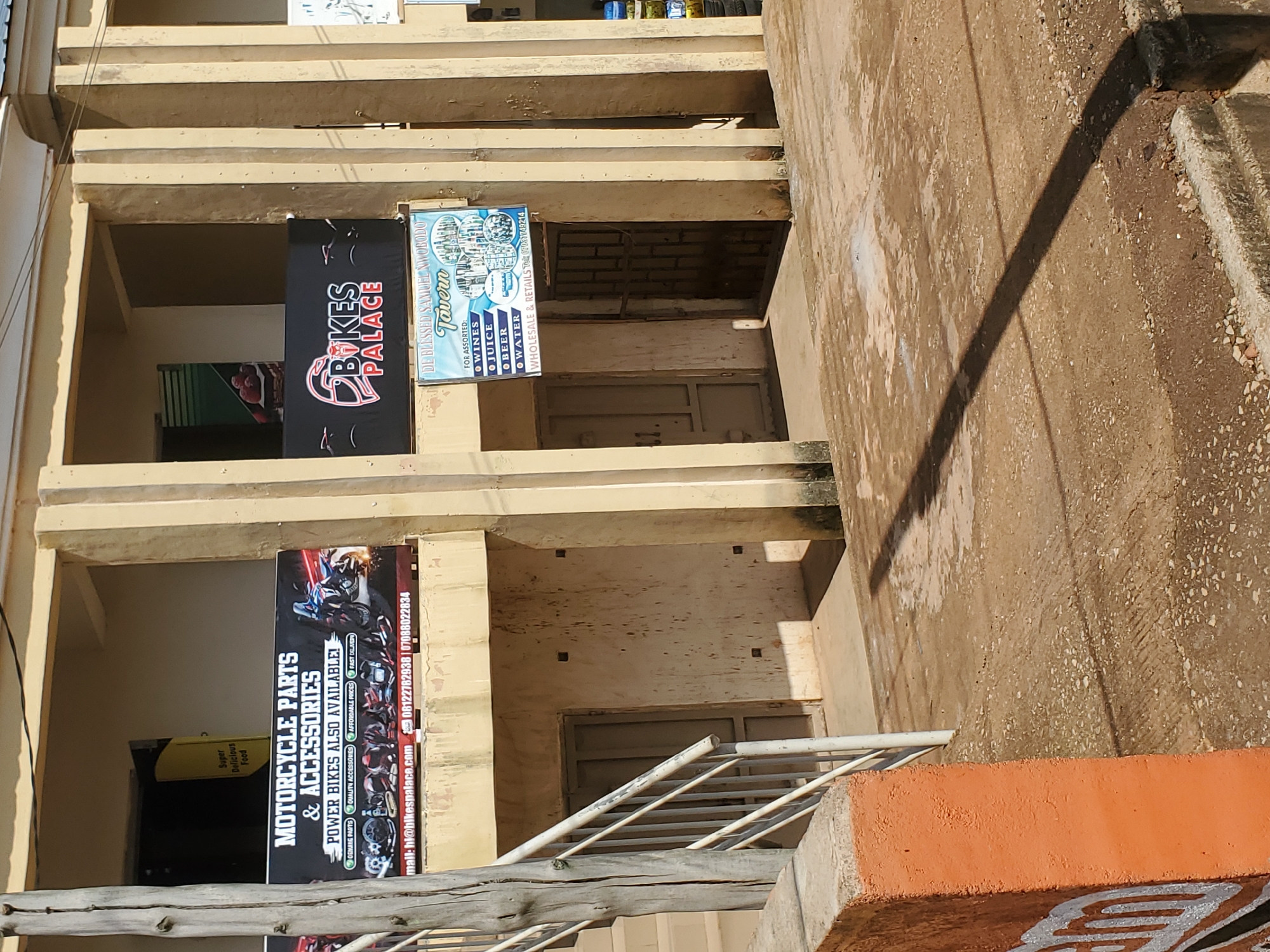 The image shows the exterior of a commercial building in Bukuru, Nigeria, with multiple signs advertising businesses. A prominent sign on the left reads "MOTORCYCLE PARTS & ACCESSORIES" with "POWER BIKES ALSO AVAILABLE!" beneath it, along with a website and phone numbers. To the right, another sign displays "BIKES PALACE" in large lettering, featuring a motorcycle helmet logo. Below that, a smaller sign advertises "DE BLESSED SAMUEL NWOBODO Tavern" offering "FOR ASSORTED: WINES, JUICE, BEER, WATER" and mentioning "WHOLESALE & RETAILS" with a phone number. The building has a beige facade with visible vertical columns. A staircase with a railing is visible on the left, leading up to the building's entrance. The ground in front of the building is paved with concrete and shows signs of wear and dirt. The overall environment appears to be a commercial street or market area.