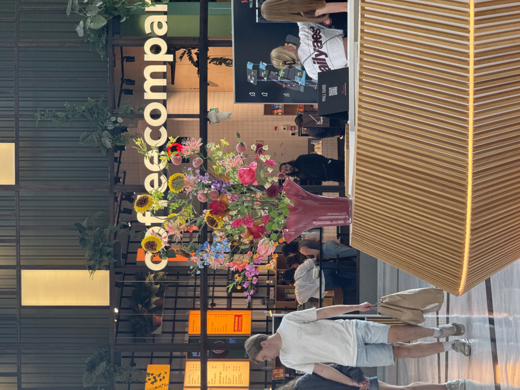 The image shows an indoor scene, likely a mall, in Leidschendam, Netherlands. In the foreground, a large, vibrant bouquet of artificial flowers is displayed in a deep red vase on a slatted wooden counter. The counter has an illuminated band of light beneath it. To the left, a young man wearing a white t-shirt and light blue shorts walks past, carrying two brown paper shopping bags. Behind him, a person's legs and lower torso are visible, also walking. In the midground, several people are visible interacting at a service counter or cafe. A large black digital screen displaying a mall map is visible behind them, with the text "MALL GUIDE" below it, and the Westfield logo. The background features a sign that reads "Coffeecompany" in large, illuminated letters, with hanging plants and shelving units visible behind it, suggesting a retail or food establishment. The overall lighting is a mix of ambient interior light and the illumination from the signs and the counter.