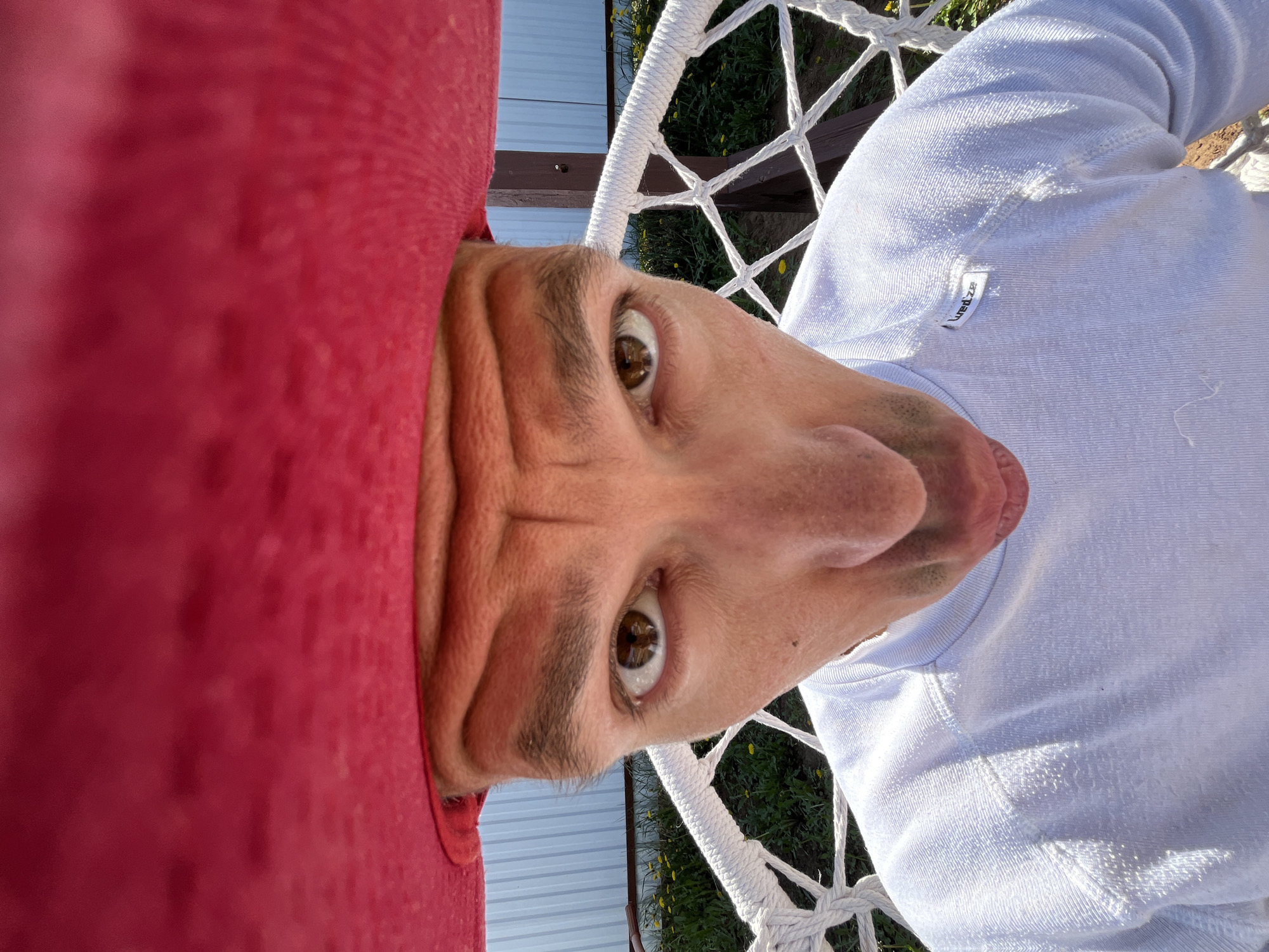 A man's face is upside-down and distorted in the image, suggesting the photo was taken from an unusual angle. He's wearing a white sweatshirt, and a red garment is visible on the left side of the frame. The subject appears to be sticking out his tongue. A white macrame hammock is behind him, and glimpses of a fence and greenery are visible in the background. No location details pertaining to the city of Chaikovsky, Russia are discernible from the image.