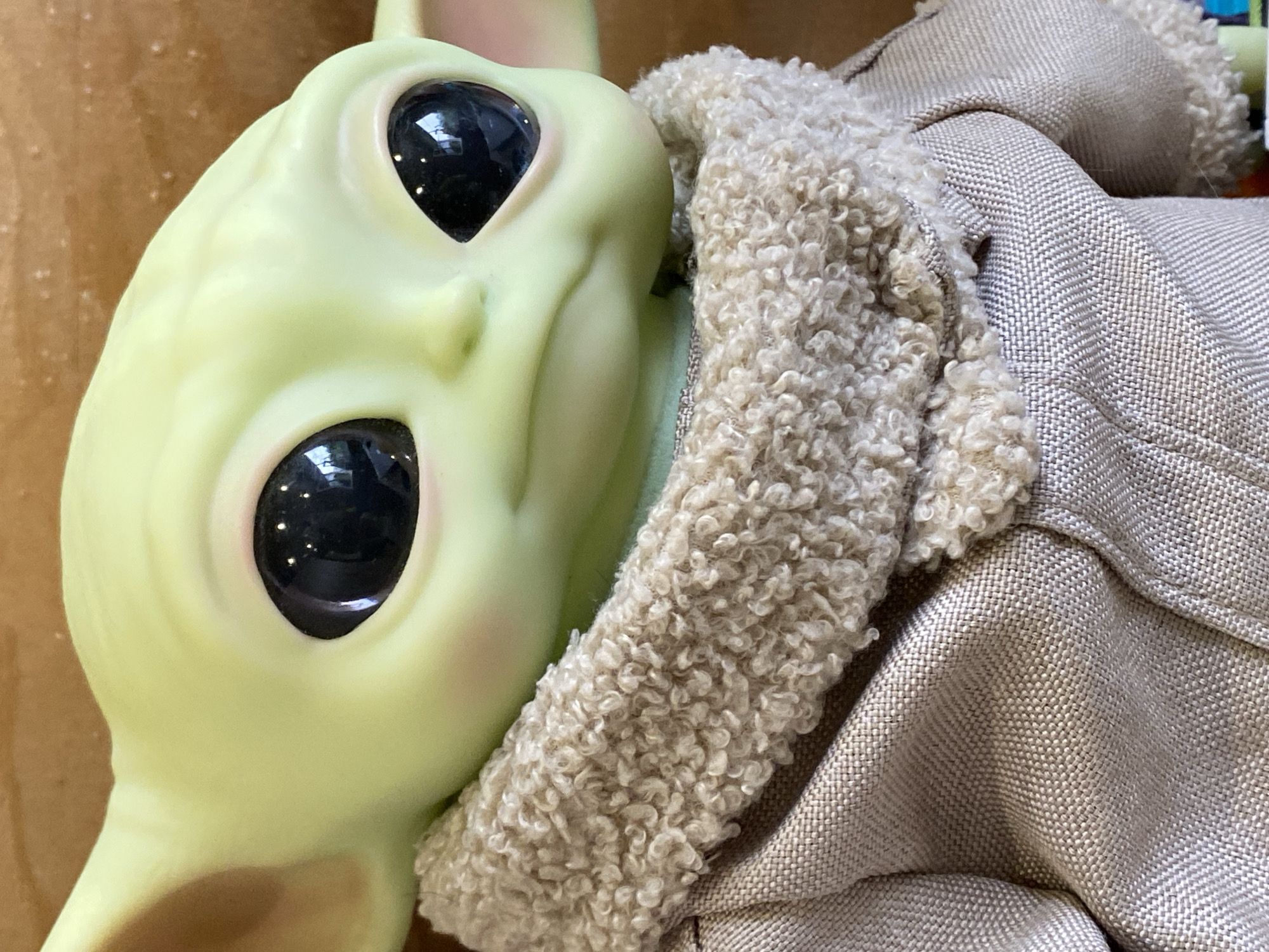 Close-up of a Baby Yoda toy, featuring a green head with large, black eyes, pink cheeks and ears, and a slight smile. The head is angled slightly. The body of the toy appears to be clad in a beige, coarse fabric coat with a fleece-like trim around the neck area. The background is a light brown color. This image was taken in a location that is not identifiable as the city of Chaikovsky, Russia.