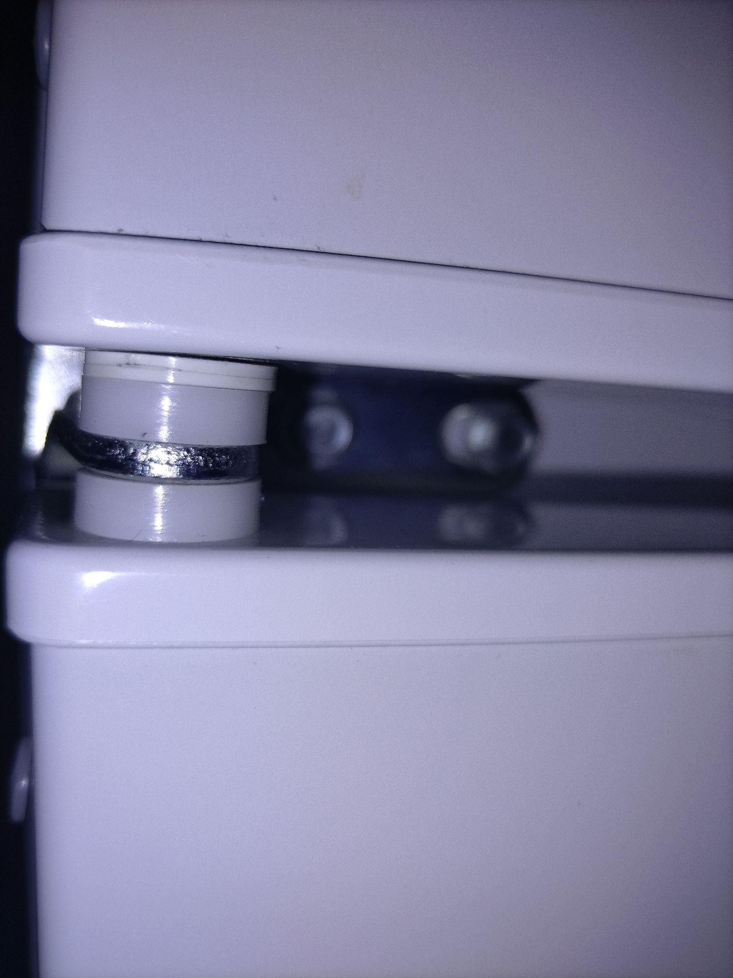 The image shows a close-up, overhead view of a white appliance with a narrow gap between two white sections. Within this gap, a cylindrical white plastic component is visible, and it appears to be a part of a locking mechanism or a hinge. A silver-colored metal ring, possibly a washer or a clip, is wrapped around the middle of the plastic component. In the background, a dark, blurry object, likely another part of the appliance or its surroundings, is visible. The lighting is somewhat dim, with reflections visible on the glossy white surfaces. The image is static and captures a small, isolated detail of the appliance. The location is described as Unknown, Unknown.