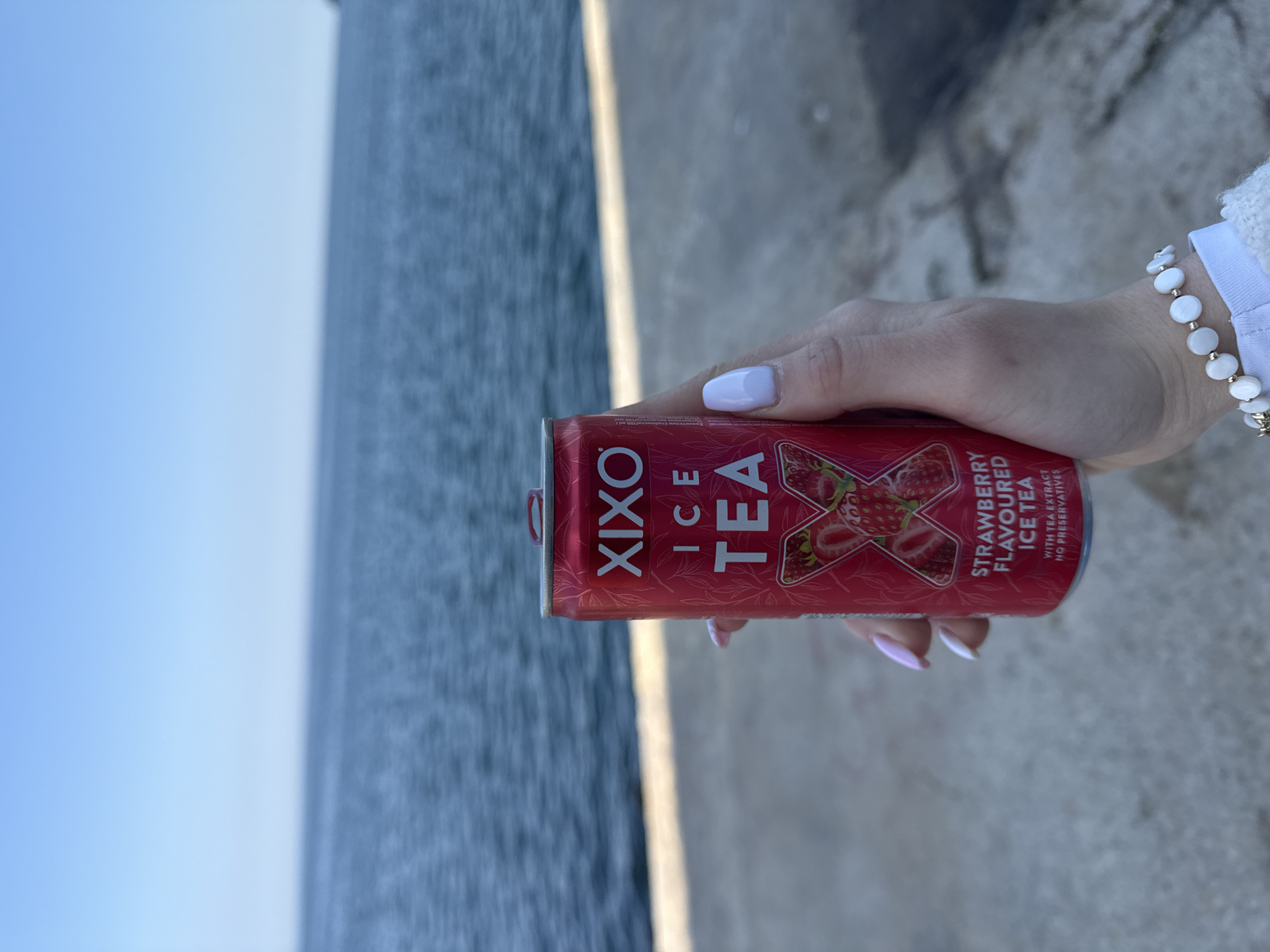 A red can of Xixo strawberry-flavored ice tea is held in a person's hand, with a portion of the Varna shoreline visible in the background. The sky is blue, and the water appears choppy. The person is wearing a white bracelet. The person's fingernails are painted white with light pink tips. The background appears to be a concrete path.