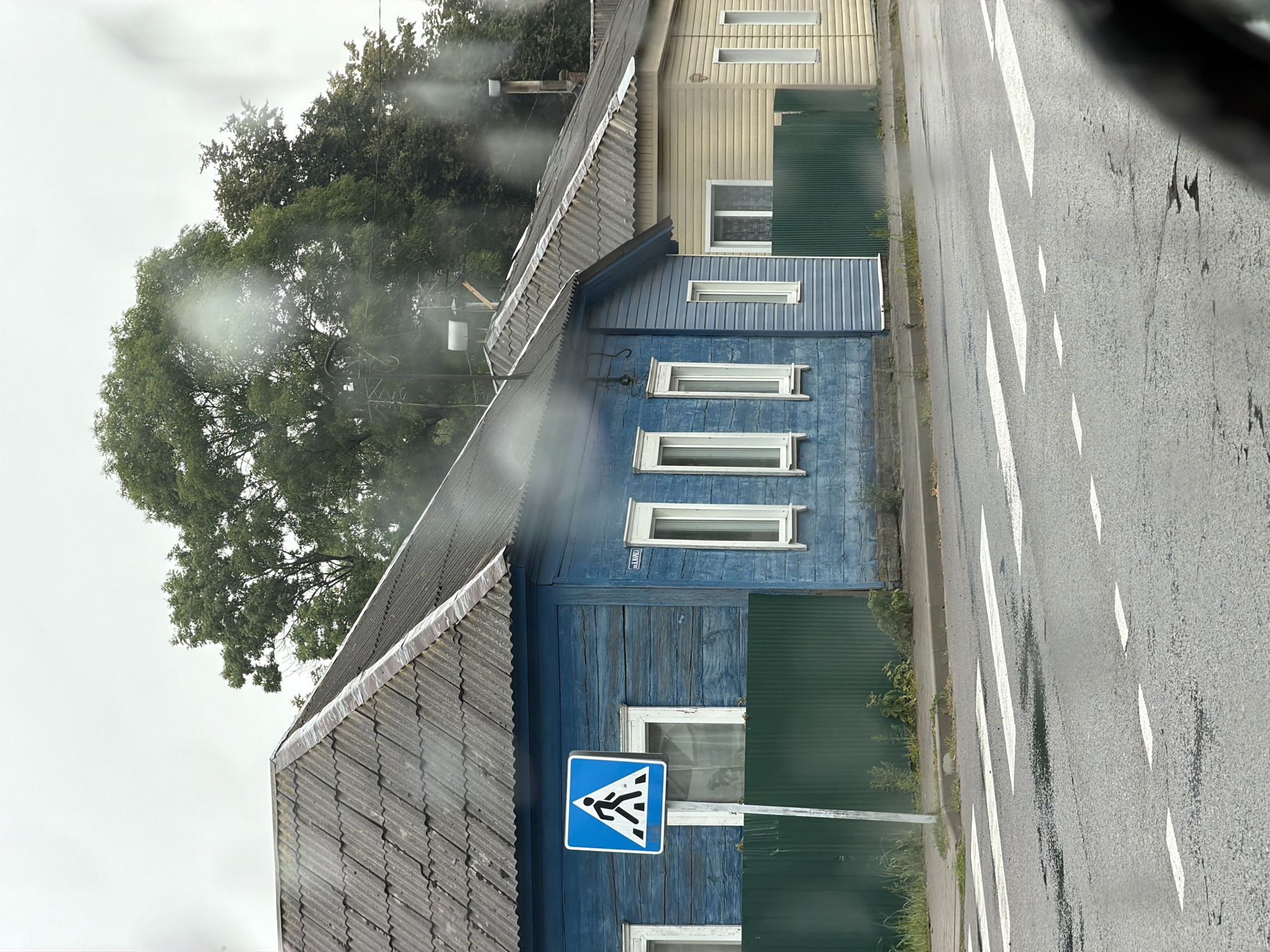 The image shows a street scene in the city of Cherven, Belarus, featuring a pedestrian crossing in the foreground and two adjacent houses. The house on the left is a blue wooden structure with a corrugated gray roof, and it has a pedestrian crossing sign in front of it. The house on the right is a lighter-colored, possibly vinyl-sided structure, also with a gray corrugated roof. Both houses have white-trimmed windows. A green metal fence separates the properties from the sidewalk and street. The road surface is asphalt and appears wet, with white lines marking the pedestrian crossing. Overcast skies are visible in the background, with trees behind the houses. The scene appears to be a residential street.