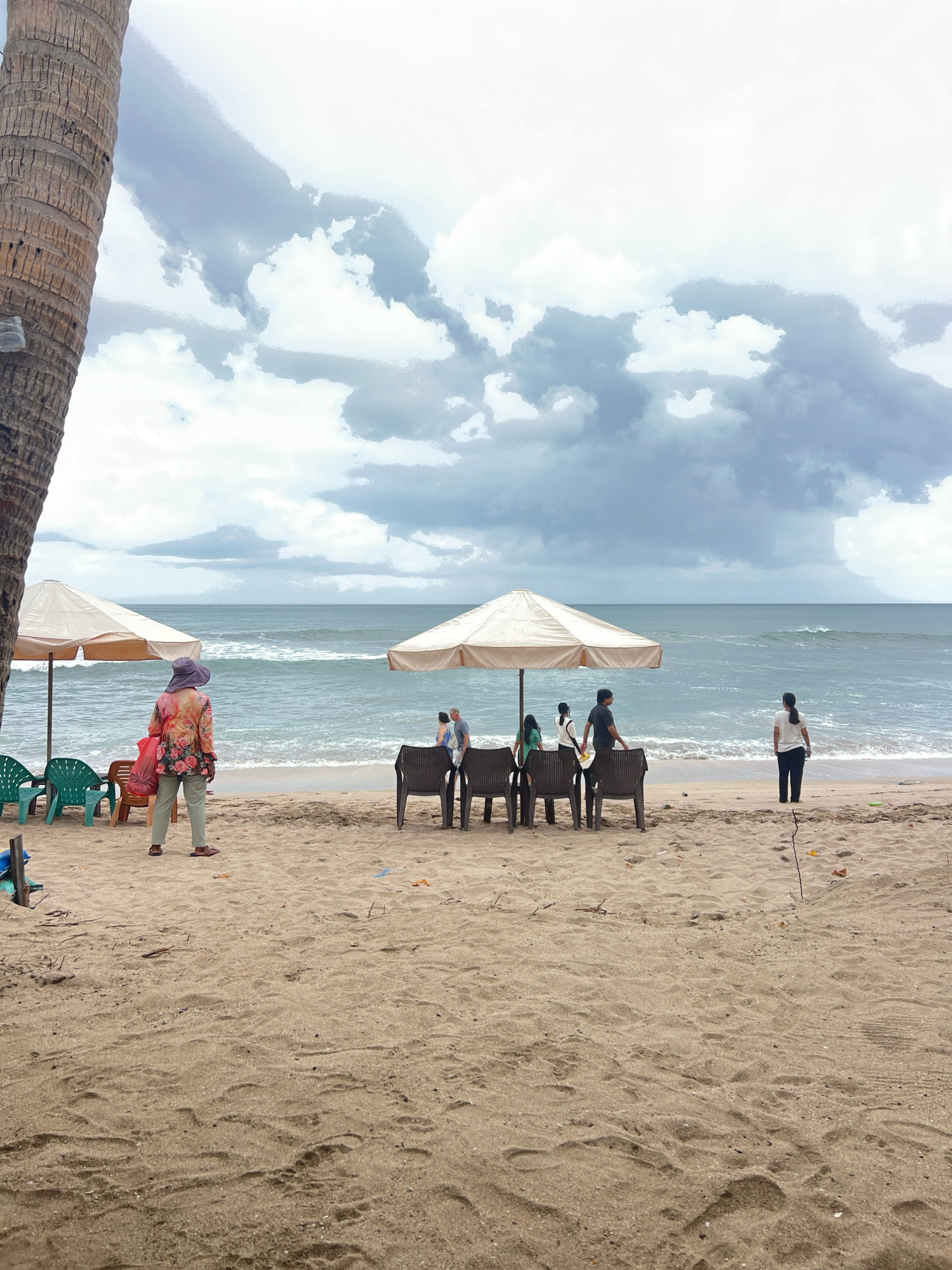 A beach scene in Kuta, Indonesia, shows a sandy shore with several individuals and beach amenities under a cloudy sky. A large palm tree trunk is visible on the left. Two beige beach umbrellas are set up on the sand, with a line of plastic chairs beneath each. Several people are present, including a woman in a floral shirt and wide-brimmed hat standing in the foreground, facing away from the camera. Near one umbrella, a group of four people are gathered, and another woman stands alone near the water's edge. The ocean, with moderate waves, stretches to the horizon, meeting the cloudy sky.
