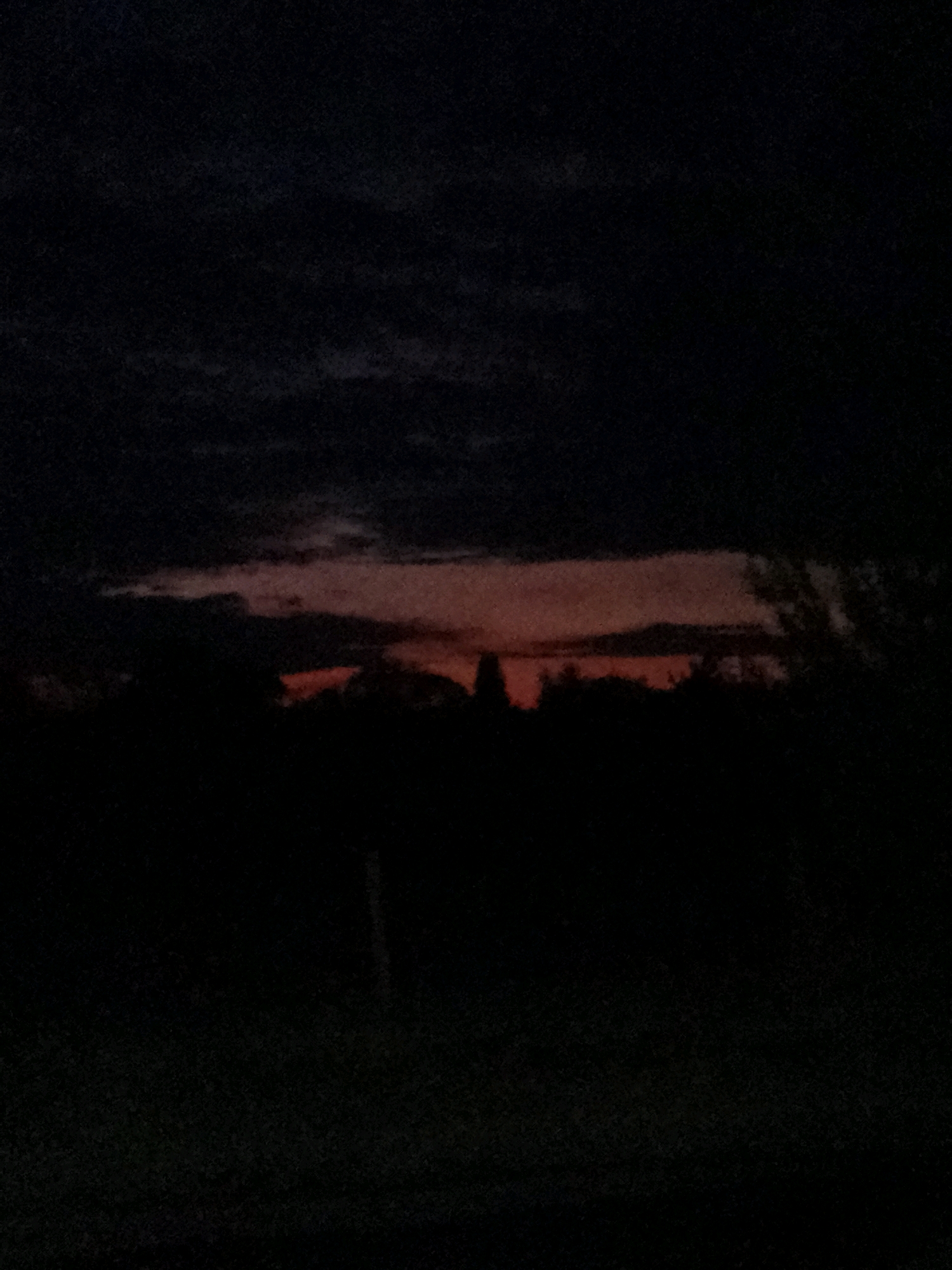 The image depicts a dark, silhouetted landscape under a twilight sky. The horizon is dominated by the dark shapes of trees and possibly other vegetation. A strip of orange and red light, indicative of a sunset or sunrise, is visible above the treeline, breaking through the otherwise dark sky. The sky above this light is a deep, dark blue or black, with faint cloud formations visible. The foreground is almost entirely black, with some subtle textures suggesting grass or uneven ground. There is a vertical dark object in the middle ground, possibly a fence post or a thin tree trunk. The overall impression is one of a rural or semi-rural setting at dusk. The location is identified as Ustyvytsia, Ukraine.