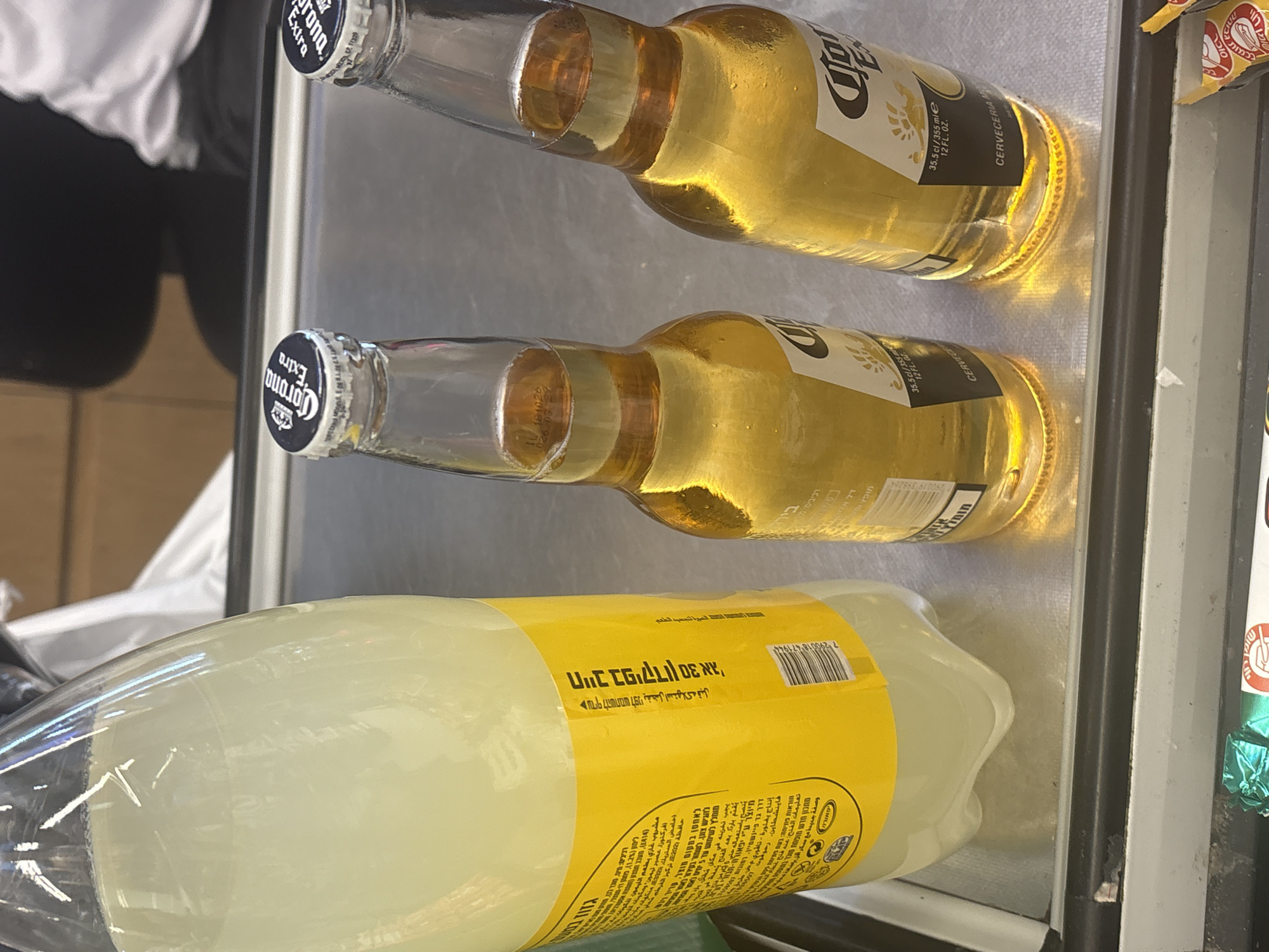This image captures a close-up view of two bottles of Corona Extra beer and a large bottle of what appears to be lemonade or a similar light-colored beverage, all placed on a metallic surface, likely a checkout counter or display shelf. The bottles are positioned in a way that highlights their labels and caps. The Corona Extra bottles, with their distinctive clear glass and golden liquid, are placed closer to the foreground, with their caps bearing the "Corona Extra" logo. The larger bottle on the left has a prominent yellow label with text in what appears to be Hebrew, indicating its origin or distribution in Israel. The background is blurred, but suggests an indoor setting with hints of human presence, possibly a store or supermarket in Tel-Aviv, Israel, given the Hebrew text. The lighting is somewhat bright, reflecting off the glass surfaces. There are no people clearly visible in the frame, but the context implies a commercial environment. The scene is static, with no discernible action taking place.