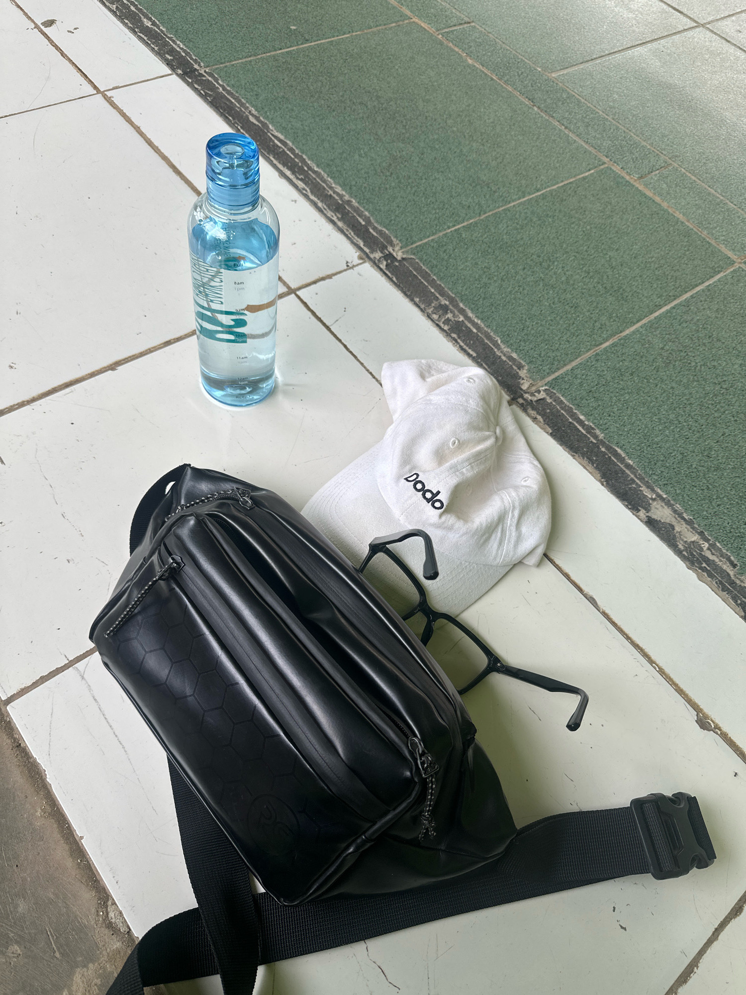 A high-angle shot shows a black fanny pack, a white baseball cap with the word "dodo" in black lettering, a pair of black-rimmed glasses, and a clear plastic water bottle with a blue cap, all resting on a tiled floor. The tiles are a light, off-white color with visible grout lines. A section of darker green tiles with a rough texture is visible in the upper right quadrant of the image, separated from the lighter tiles by a darker strip of material. The fanny pack is positioned in the foreground, with its strap extending to the bottom right. The cap is placed on top of the fanny pack, and the glasses are positioned next to the cap. The water bottle stands upright to the left of the cap. The overall scene is set outdoors, possibly on a walkway or patio, given the tiled surface. There are no people visible in the image.