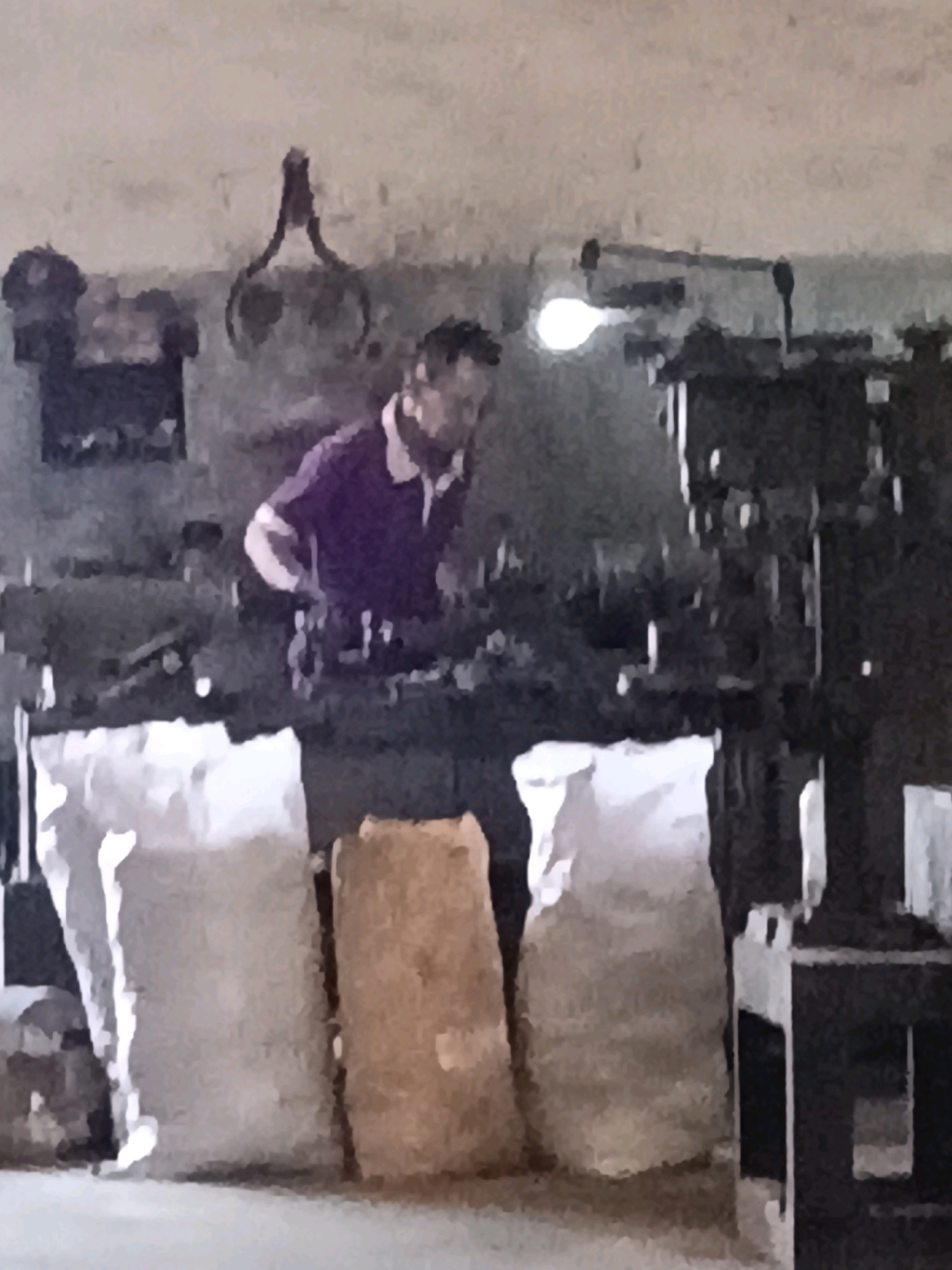 The image depicts an indoor scene, possibly a workshop or factory, in the city of Belimbing, Indonesia. In the center of the frame, a person wearing a purple polo shirt is visible, appearing to be engaged in some form of work. The person is in focus, while the surroundings are somewhat blurred. To the left of the person, there are three large sacks on the floor, two of which are white and one appears to be made of burlap or a similar material. Behind the person, there are industrial machines and equipment, including a bright light source overhead, suggesting an operational environment. The walls of the room are plain and appear to be made of concrete or plaster. The overall impression is of a functional workspace where production or manufacturing activities are taking place.