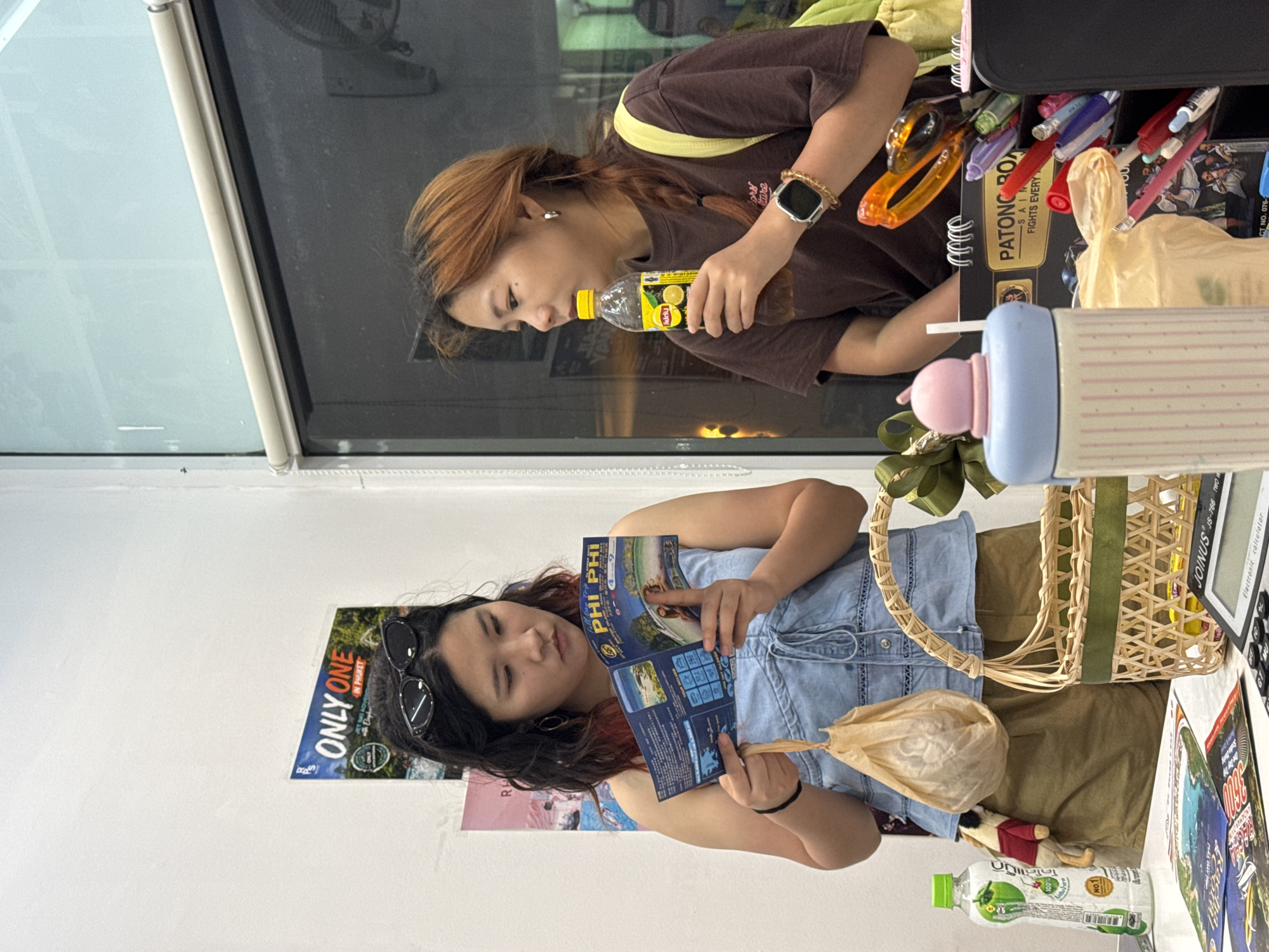 This media file features two individuals in what appears to be an indoor setting, possibly a shop or a waiting area in Karon, Thailand. The scene is captured in a slightly tilted, vertical orientation.
In the foreground, a person with dark hair, sunglasses perched on their head, and wearing a denim vest over a blue top is holding and examining a brochure. The brochure appears to be a travel guide for "Phi Phi Islands." They are also holding a small, light-brown paper bag. Visible text on the wall behind them includes "ONLY ONE IN PHUKET."
In the background, another person with reddish-brown hair, wearing a brown t-shirt and a yellow strap across their shoulder, is holding and drinking from a bottle of Lipton iced tea. They are wearing a smartwatch on their left wrist. On a desk or counter behind them, there are various items, including pens, scissors, and a notebook with text that includes "PATONG ROAD" and "FIGHTS EVERY".
The lighting suggests it could be daytime, with natural light coming from a window or a bright overhead source. The overall mood seems relaxed, with both individuals engaged in their respective activities. The presence of travel-related materials and brand names strongly suggests a tourist-oriented location.