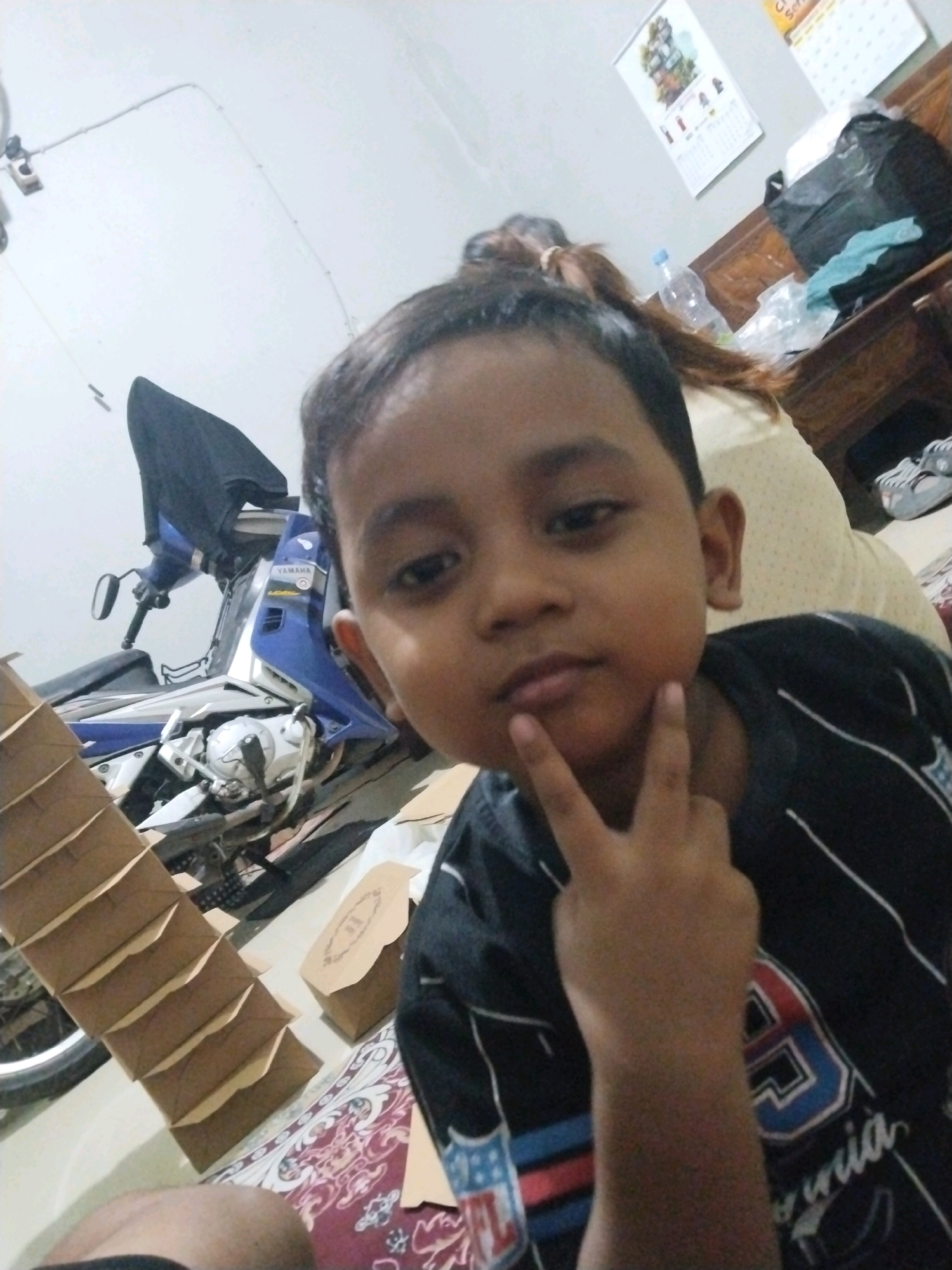 In Pepelegi, Indonesia, a young boy with dark hair and brown eyes is pictured in a close-up shot. He is wearing a dark t-shirt with white and red stripes and a graphic design featuring the number 39 and the word "win". The boy is holding up two fingers in a peace sign near his chin. In the background, a blue and silver Yamaha motorcycle is visible, covered with a black cloth. To the left, a stack of brown cardboard boxes is arranged in a staircase formation. Further back, on a wooden table, there are calendars, a plastic water bottle, a black trash bag, and a pair of grey sneakers. The environment appears to be indoors, possibly a home or a workshop, with a light-colored wall and a patterned rug on the floor.
