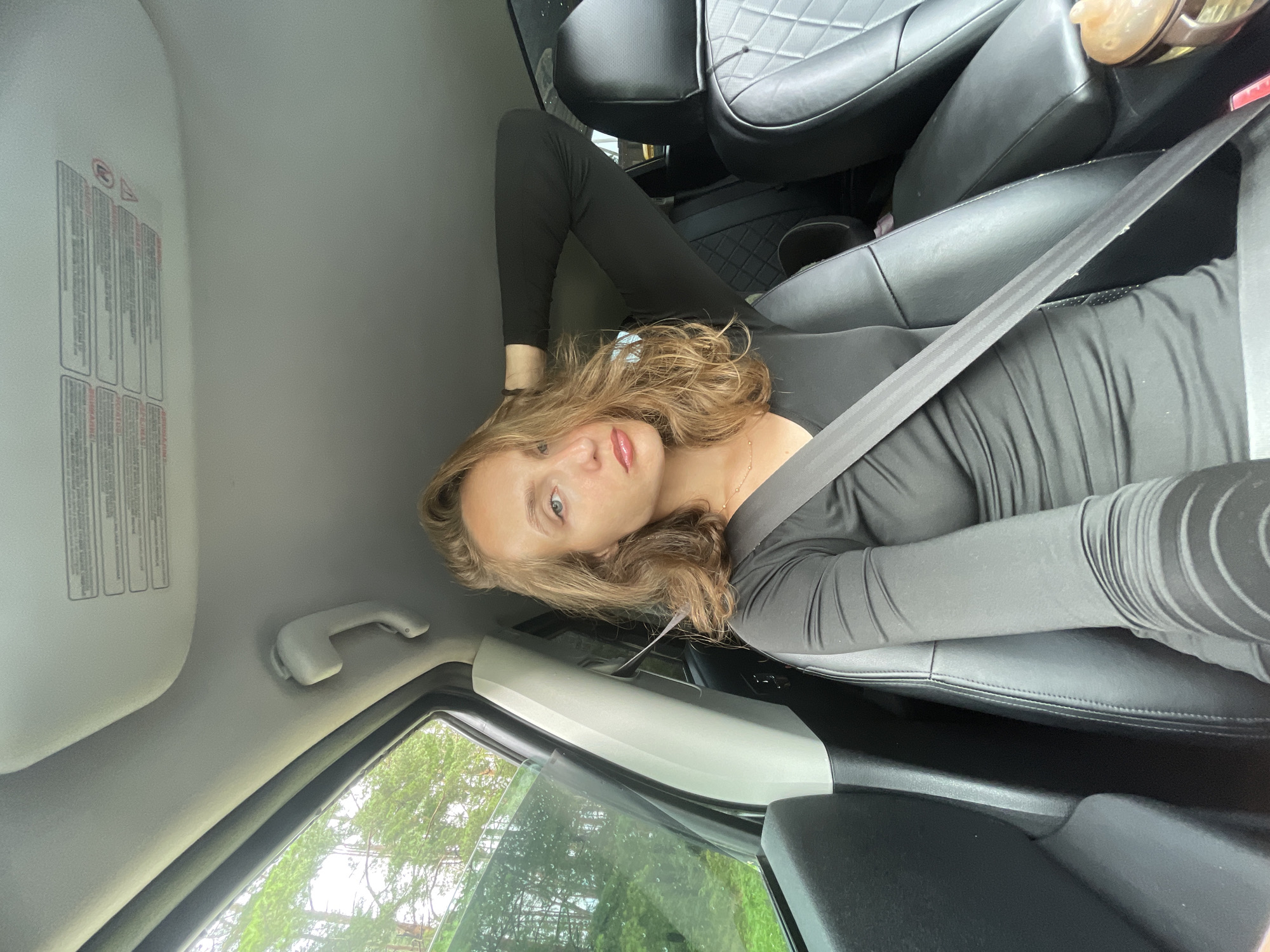 A woman with blonde hair is sitting in the driver's seat of a car. She is wearing a black long-sleeved shirt and a grey seatbelt. Her left arm is raised, with her hand on the back of her head. The car's interior is visible, including black leather seats. Outside the car window, trees and greenery are visible, suggesting a rural or wooded setting. The sky appears overcast. There are safety labels visible on the sun visor. The location is identified as Podshivalovo, Russia.