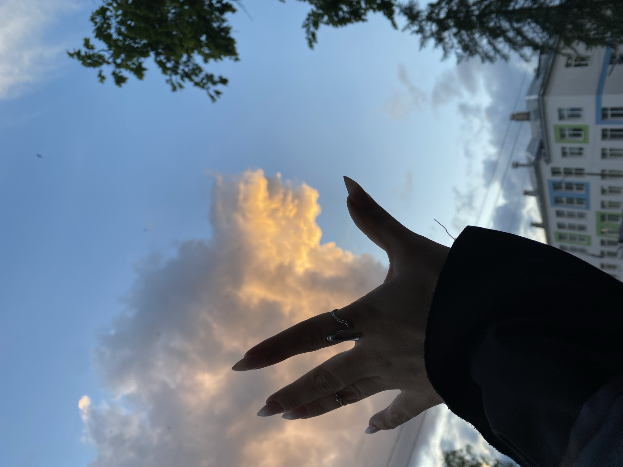 The image depicts a low-angle view of a hand extended towards the sky, with clouds illuminated by the setting sun. A building is visible in the background, along with tree branches. The hand has two rings, one on the index finger and one on the ring finger. The location of the image is in the city of Chaikovsky, Russia.