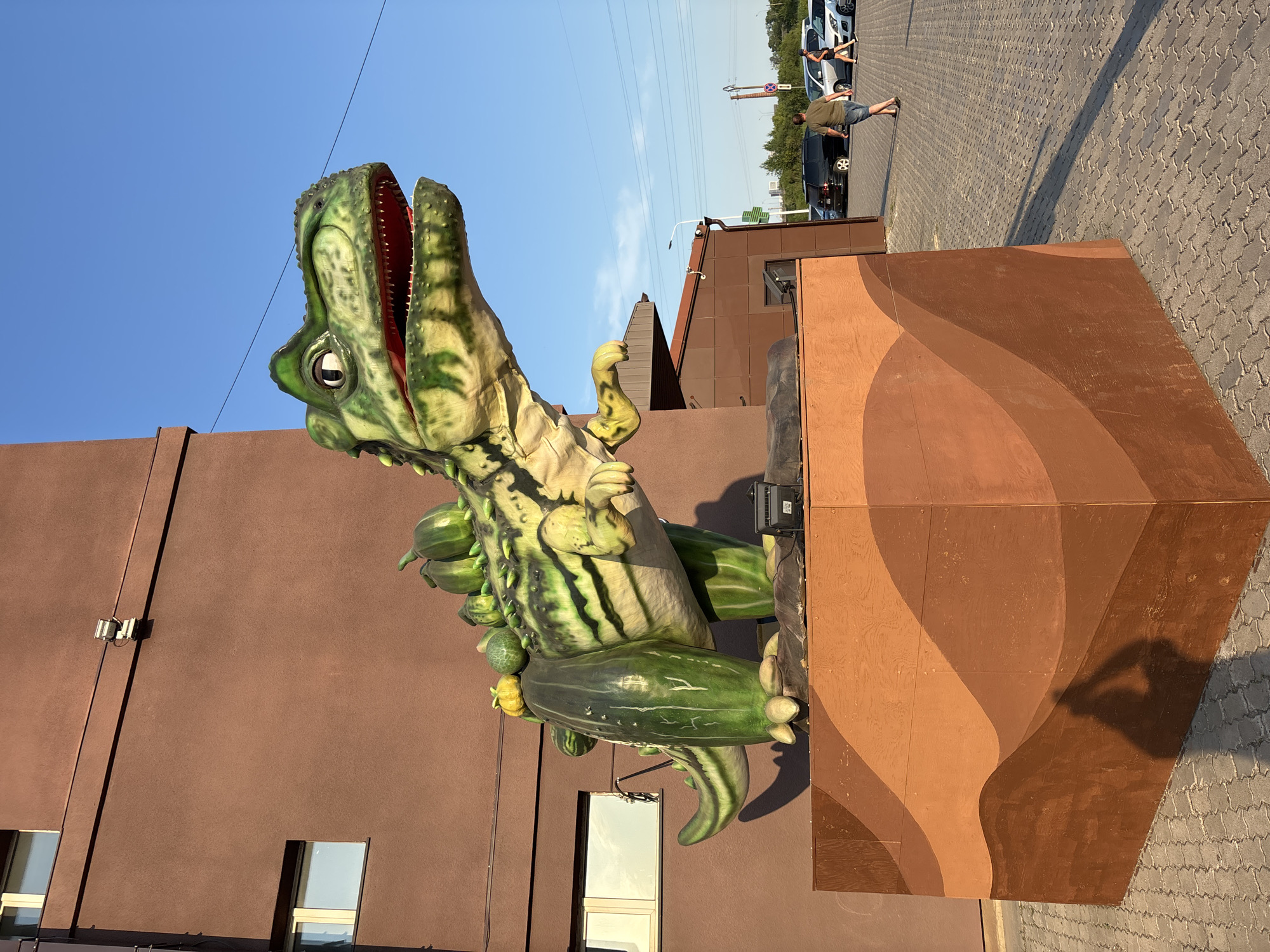 This media file depicts an outdoor scene, likely during the daytime under a clear blue sky. The primary subject is a large, green, dinosaur-like sculpture with its mouth wide open. The sculpture appears to be made of fiberglass or a similar material and is painted with darker green and yellow markings. It is positioned partially emerging from a brown, abstractly shaped structure.
In the background, a building with a brown facade and several windows is visible. To the right of the dinosaur sculpture, a paved area with a repeating pattern of rectangular stones is present. On this paved area, there are a few people and cars, suggesting a public space such as a parking lot or an outdoor entertainment venue. One person is walking, and another is sitting or leaning against a car. The sunlight casts long shadows, indicating it might be late afternoon or early morning.
The overall scene conveys a playful or perhaps kitschy atmosphere, with the exaggerated dinosaur sculpture being the focal point. There are no visible signs or text in the image. The location context provided is Blagoveshchensk, Russia, which might explain the architectural style of the building or the presence of certain vehicles, though these are not definitively identifiable from the image alone. The weather appears to be fair and sunny.