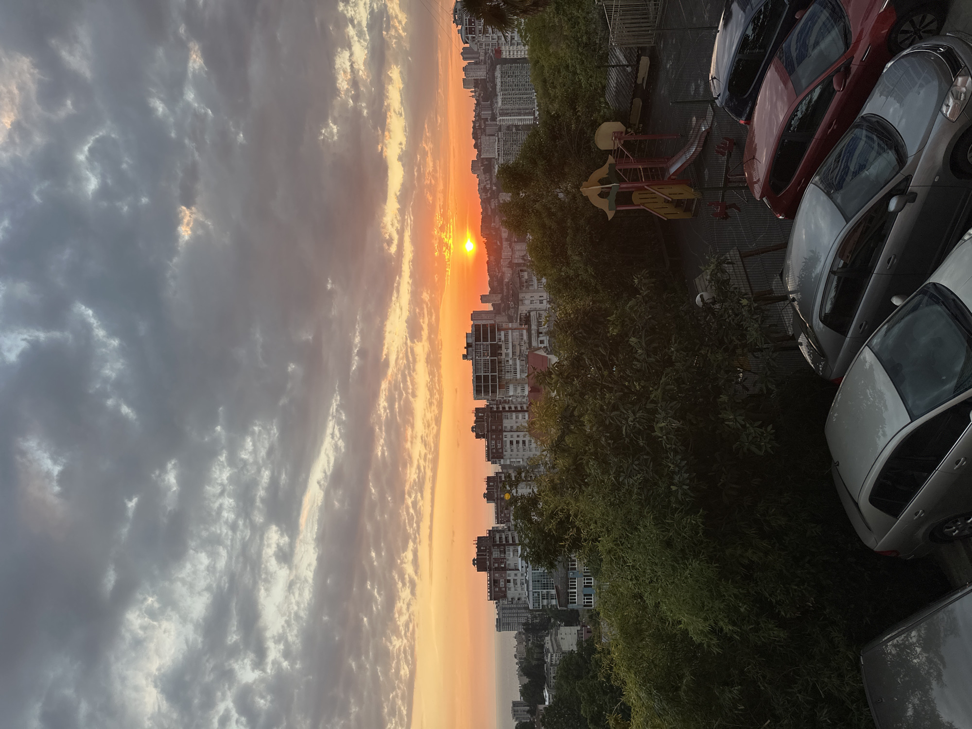 This is an outdoor, medium-shot, vertical-format, eye-level view of a city skyline at sunset, likely in Sochi, Russia. The sun, a bright orange orb, is low on the horizon, casting a warm glow across the sky. The sky is filled with dramatic, layered clouds ranging from light gray to deep charcoal, with streaks of orange and pink where the sun's rays penetrate. In the middle ground, a dense cluster of multi-story buildings, many with red-tiled roofs, dominates the urban landscape. Some buildings have visible balconies and windows. Lush green trees and foliage are prominent in the foreground and middle ground, partially obscuring the view of the buildings and a children's playground with a slide and swings. Several cars are parked in a lot in the lower right of the image, including a dark SUV, a red hatchback, and two silver sedans. The ocean is visible in the far distance on the left, meeting the horizon. The overall mood is serene, with the natural beauty of the sunset contrasting with the urban environment.