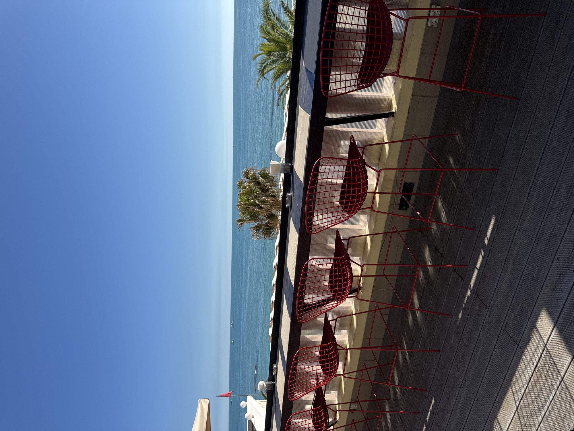 The image depicts a sun-drenched outdoor patio or balcony overlooking a calm, deep blue sea under a clear, lighter blue sky. The scene is oriented vertically, suggesting a portrait shot rotated on its side.
In the foreground, a series of red, modern, wire-framed bar stools are arranged neatly along a dark, wooden-topped bar or high table. Their intricate metal frames cast strong, geometric shadows onto the light brown wooden deck floor, indicating bright sunlight. Several small, white objects, possibly condiment holders or small lamps, are visible on the table.
Beyond the patio edge, lush green palm trees add a tropical element to the setting, separating the viewing area from the expansive sea. The sea is tranquil, with only a few tiny boats or distant vessels visible on its surface. A small red flag is also noticeable near the water's edge on the left side of the image. The sky above is a brilliant, cloudless blue, transitioning to a slightly paler hue near the horizon. Part of a light-colored umbrella or awning is visible on the far left, suggesting further outdoor seating.
The absence of people suggests a quiet moment, perhaps early morning or late afternoon. The bright lighting, clear sky, and calm sea point to excellent weather conditions. The overall atmosphere is one of serenity and relaxation, characteristic of a coastal resort setting, which aligns with the provided location context of Sochi, Russia. There is no visible text in the image.