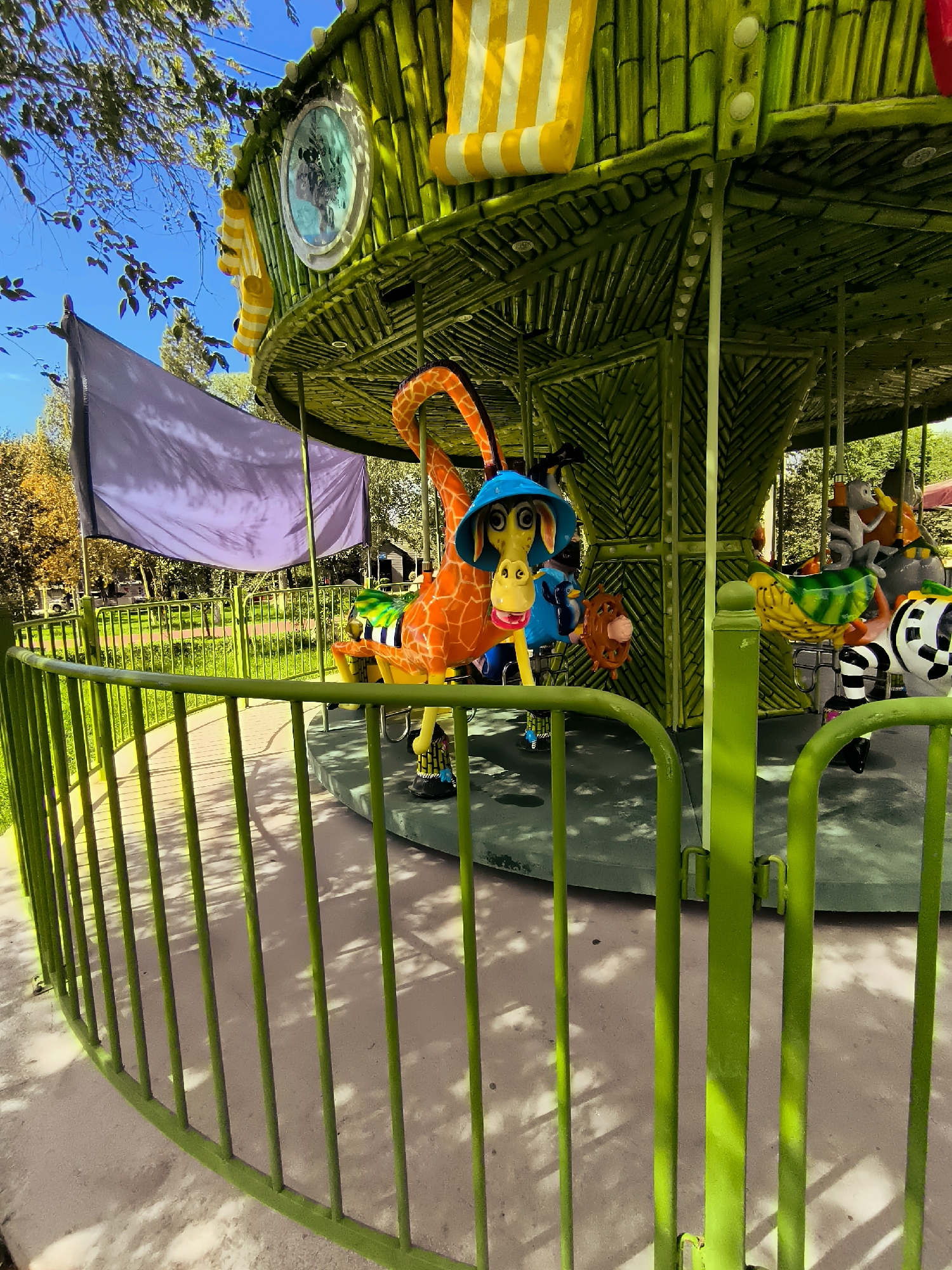 The image depicts a close-up, eye-level, outdoor, medium shot of a children's carousel. The carousel structure is primarily green and designed to resemble bamboo. It features several animal figures as seats, including a prominent orange giraffe wearing a blue hat, a zebra, a bird, and what appears to be a monkey. The carousel is surrounded by a curved, bright green metal fence. In the background, there is a large purple flag, trees with yellowing leaves, and a clear blue sky. The carousel appears to be stationary. The ground around the carousel is a light-colored paved surface with dappled shadows. The overall impression is of a playful and colorful amusement ride in a park-like setting, suggesting it is located in a city with parks and recreational facilities.