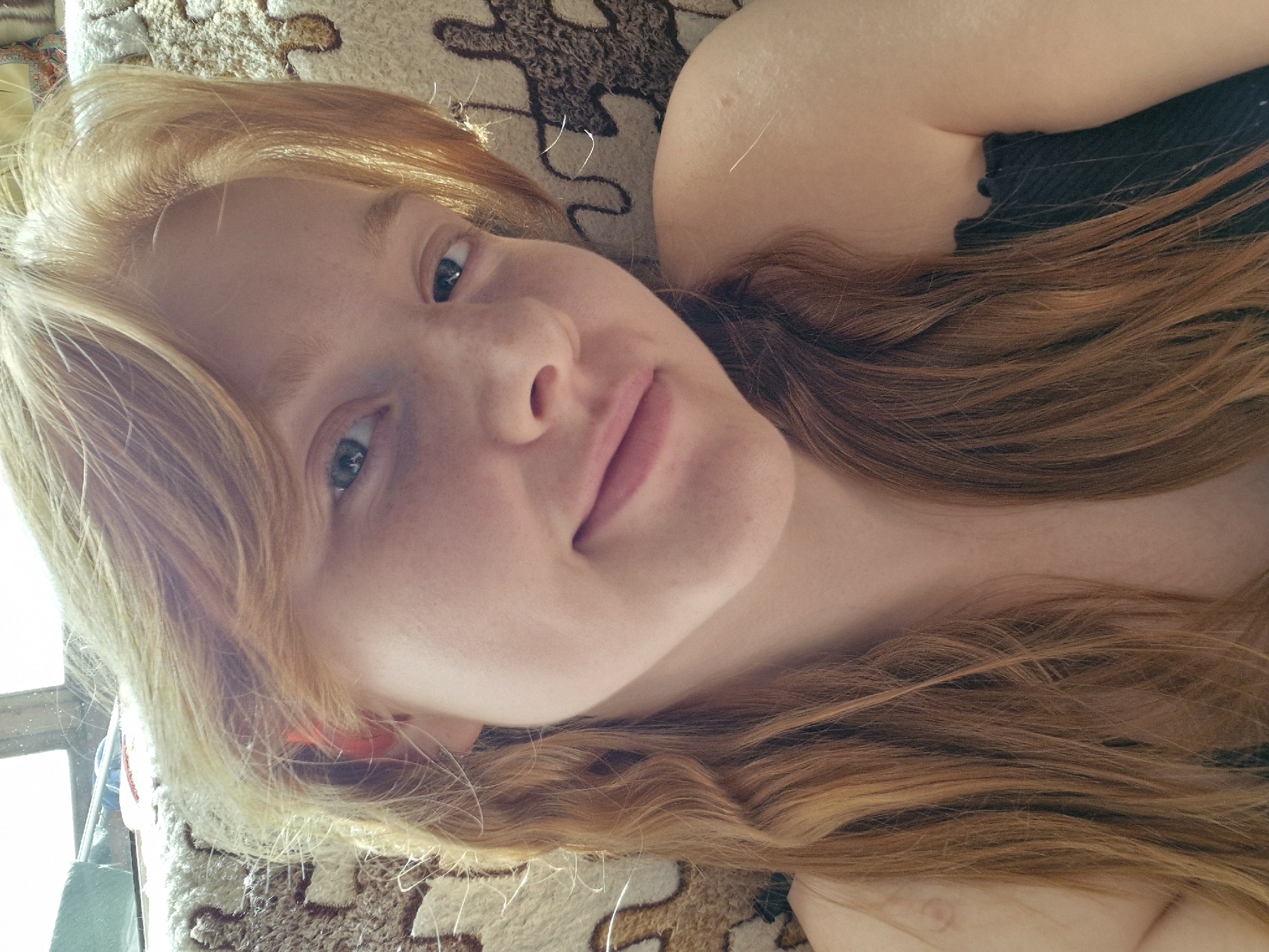 The image depicts a person lying down on a patterned surface. The person has fair skin, freckles, and reddish-blonde hair. They are wearing a dark-colored top. The location is unspecified and there is no indication that it is Yekaterinburg, Russia. The person is looking towards the camera with a slight smile. There are no other people or distinct objects present. The interaction is simply a direct gaze towards the camera. The environment is confined to the patterned surface and a glimpse of a window frame in the background.