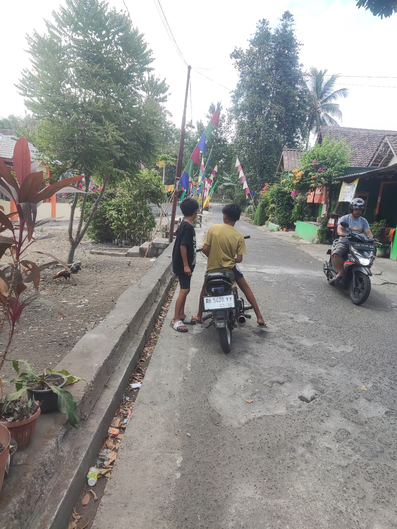 In Trihanggo, Indonesia, two boys are on a motorcycle on a paved road. One boy, wearing a yellow shirt, is seated on the motorcycle, while the other, in a black shirt, stands beside him. A man is riding another motorcycle, wearing a gray shirt and a helmet, approaching them from the other direction. The road is lined with trees and houses. Colorful flags are strung across the road. A chicken is pecking at the ground on the left side of the road.