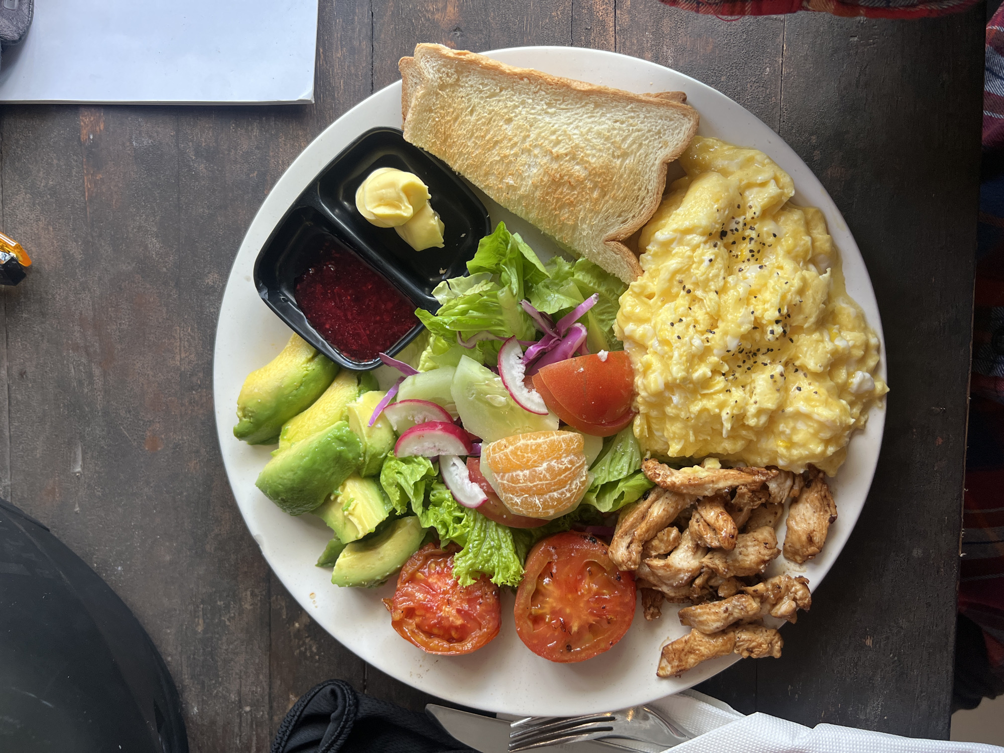 The image depicts a plate of food on a dark wooden table. The plate contains a variety of items including scrambled eggs with pepper, sliced avocado, a mixed green salad with tomato, cucumber, radish, and orange segments, grilled chicken, a slice of toast, and a small container with butter and jam. Cutlery and a napkin are visible at the bottom of the image. The setting suggests an informal dining environment in a place located in the city of Tibubeneng, Indonesia.