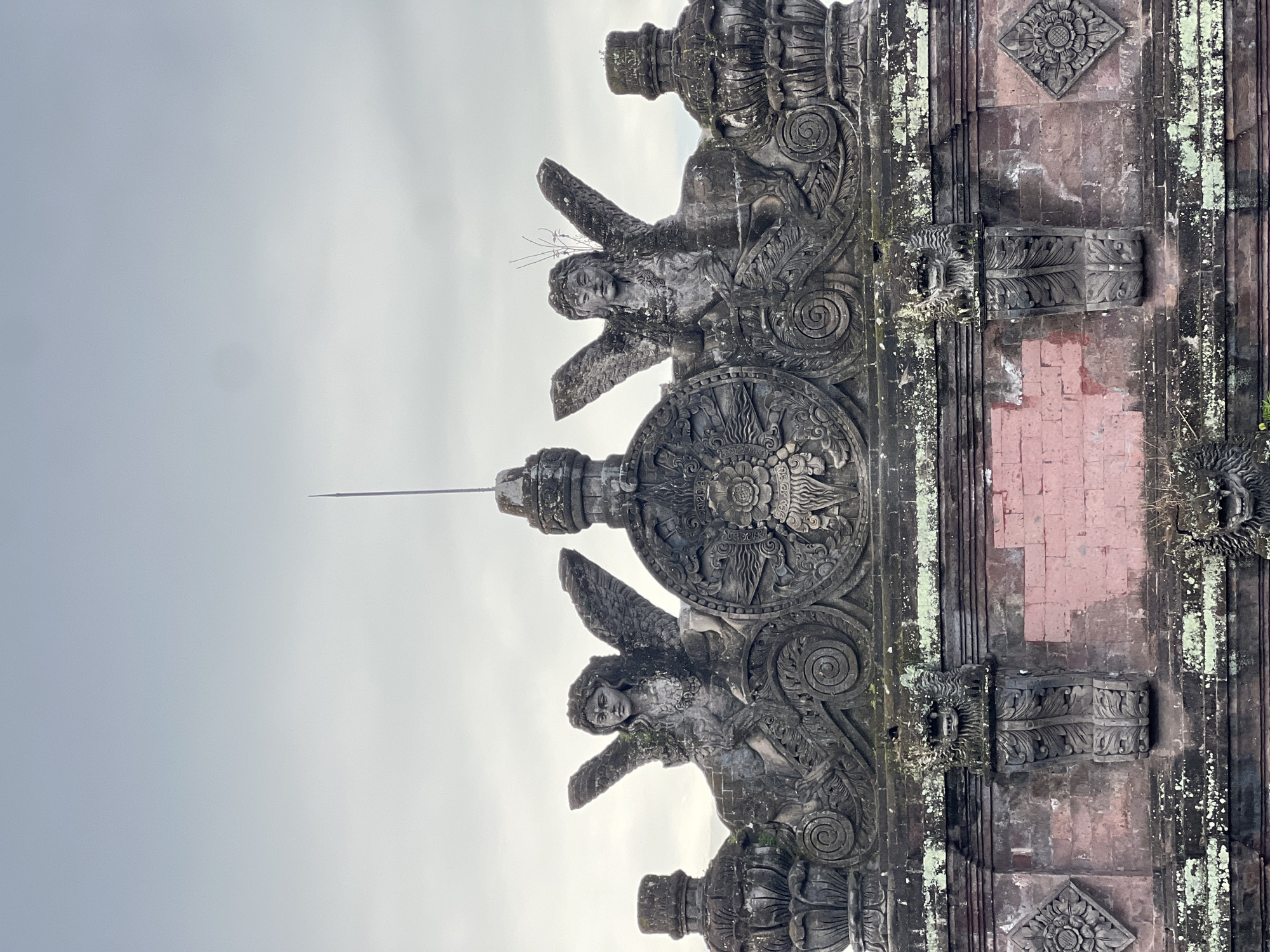 A low-angle shot captures an ornate structure, likely part of a temple or gate, in Celuk, Indonesia. The structure is predominantly stone, with intricate carvings of figures that seem to be angels or winged deities flanking a circular emblem. A tall, thin spire extends upwards from the center. The stone's color varies from dark grey to a muted red-brown, and some sections show signs of age with patches of moss or lichen. The sky is overcast and grey.