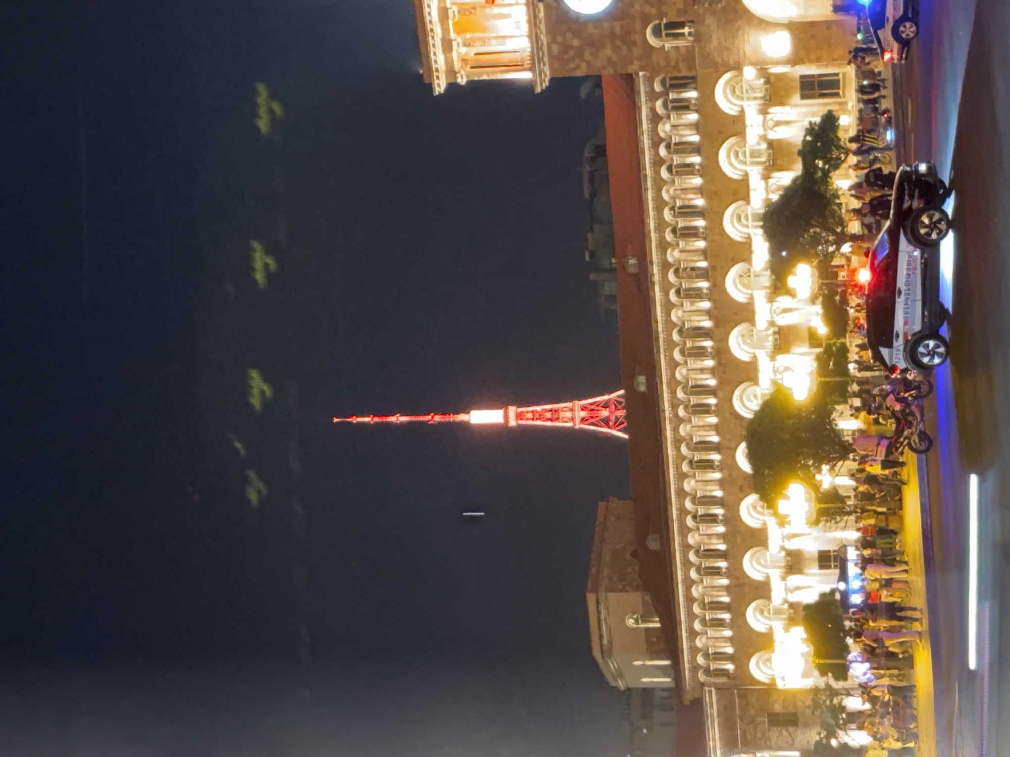 This is a nighttime photograph taken in Republic Square in Yerevan, Armenia. A tall, red and white lit tower dominates the skyline. In the foreground, a busy street scene unfolds. A police car, with its lights flashing red and blue, is stopped in the middle of the road, facing right. Another police car is visible further down the street on the right. A group of people are gathered on the sidewalk in front of a large, illuminated building with arched windows. Some individuals are walking, while others appear to be socializing. A motorcycle is parked to the right of the police car in the foreground. The overall atmosphere suggests a lively evening in the city.