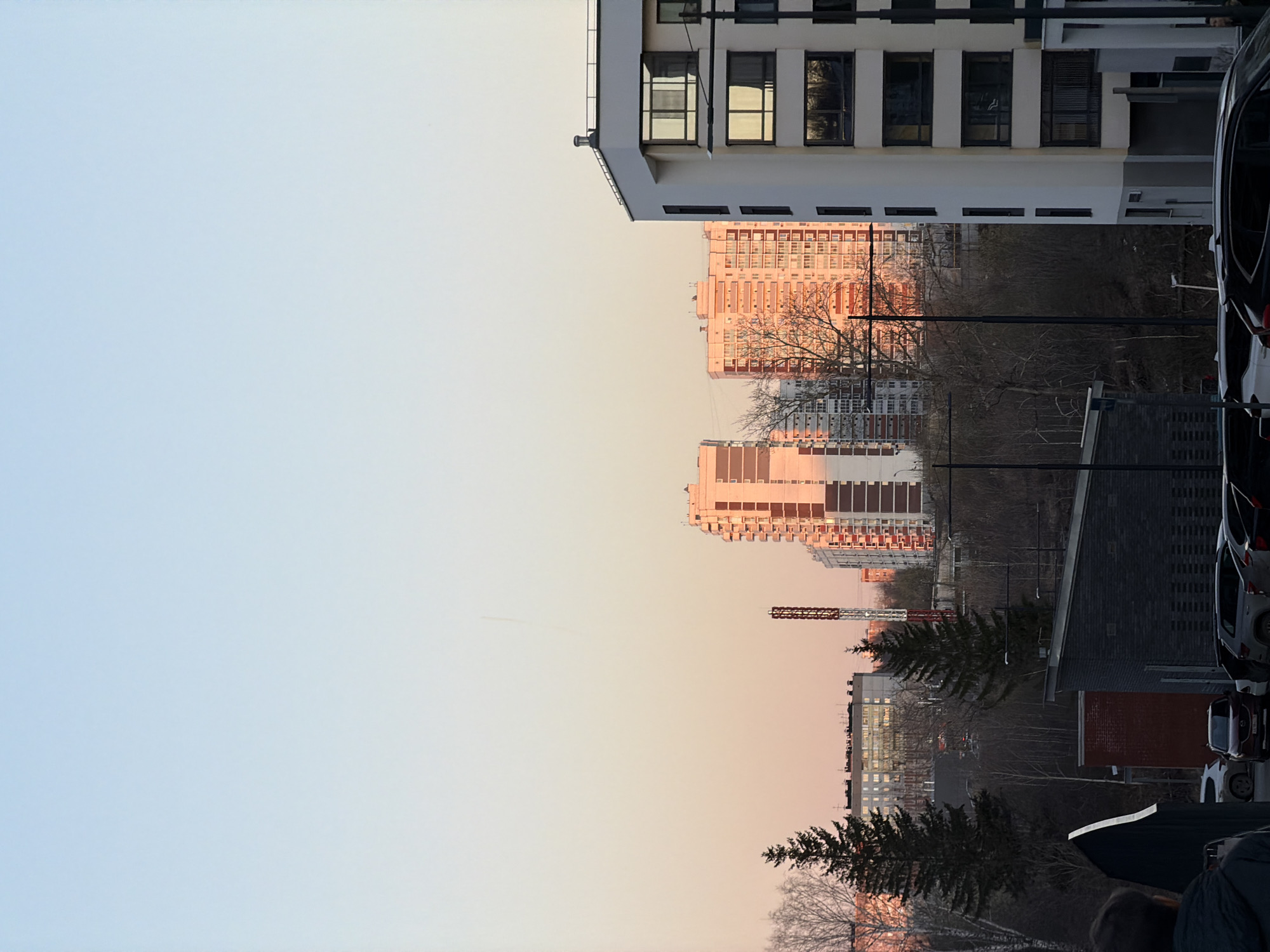 The image shows a cityscape in Izhevsk, Russia at sunset. To the left, a pale blue and pink sky takes up approximately half the frame.
On the right half of the image, buildings are visible. A modern white building with large windows is in the upper right corner. Adjacent to this, further into the distance, are orange-colored apartment buildings, presumably high-rise. There is bare tree branches between the buildings. Below this are other buildings of various heights, and a construction crane. Cars are parked along the right edge of the frame. A dark-haired person can be seen in the very bottom right corner of the frame, partially obscured.