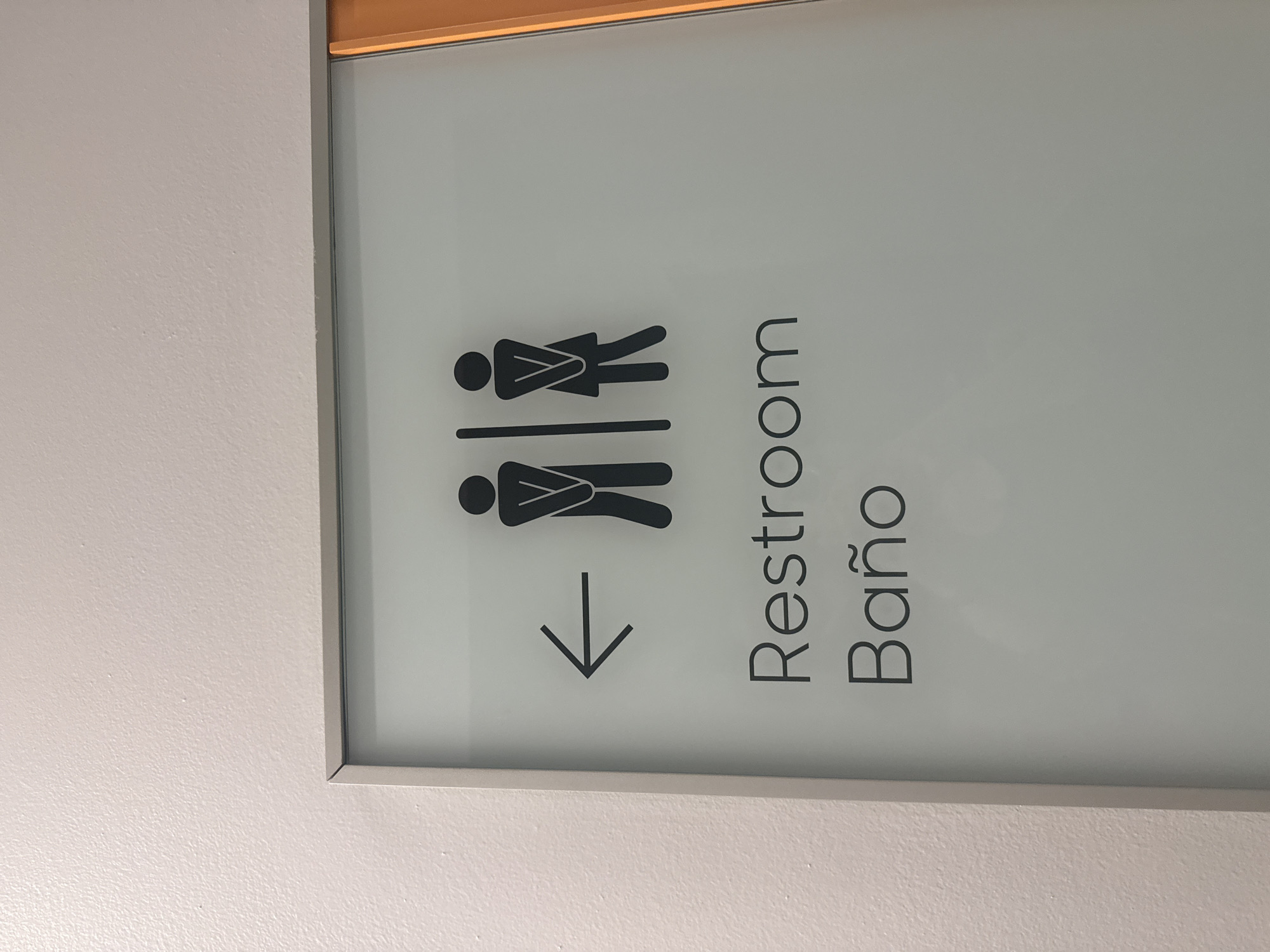 The image shows a restroom sign mounted on a white wall, indicating the direction of the restroom with an arrow pointing downwards and to the left. The sign displays pictograms representing both male and female figures, along with the words "Restroom" and "Baño" written in a simple, sans-serif font. The sign has a silver frame with a thin gold trim at the top. The location is likely a public space somewhere in the United States of America as indicated by the English word for restroom, and more precisely Miami, United States of America according to the request.