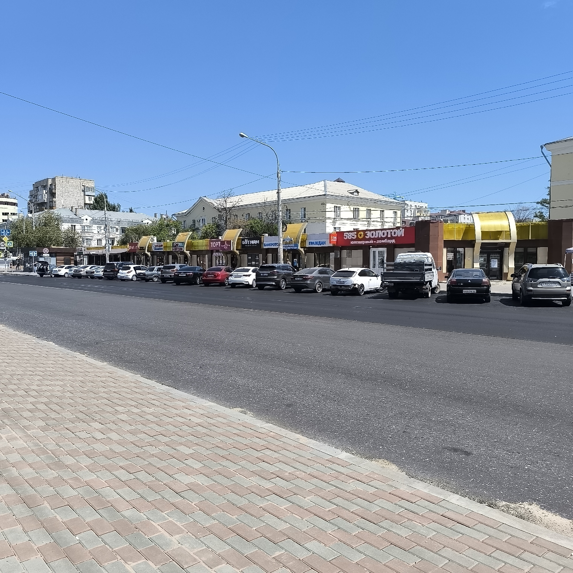 The image depicts a street scene in Astrakhan, Russia, on a clear day with a blue sky. The foreground shows a sidewalk paved with reddish-brown and gray rectangular bricks. The street itself is asphalt. On the right side of the street, a line of parked cars is visible. These include a white sedan, a dark gray sedan, a black sedan, a silver SUV, and a white hatchback. Further down the street, a white pickup truck with a black flatbed is parked.
Adjacent to the street are various commercial establishments. One building features a prominent yellow awning and signage reading "585 ЗОЛОТОЙ" (585 GOLD), indicating a jewelry store and pawnshop. Other visible store signs include "ТОРГ" (TRADE), "GÜTHAM", and "БАНКФОТО" (BANKPHOTO). Buildings appear to be of various architectural styles, with some having light-colored facades and multiple windows. Trees with green foliage are interspersed between the buildings. In the background, a multi-story residential building with a light-colored facade and numerous balconies is visible. Power lines and a streetlamp are also present in the scene. The overall impression is of a bustling urban commercial area.