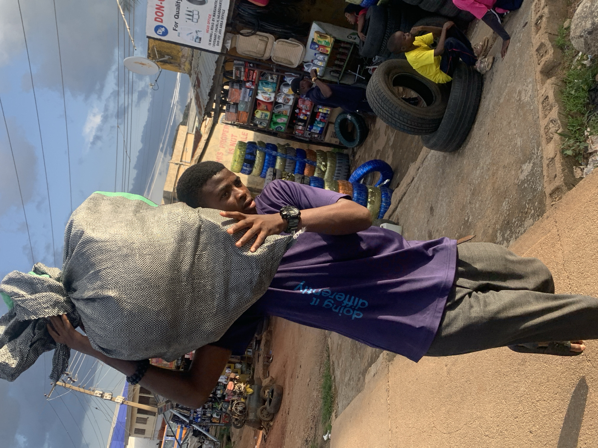 The image depicts a young man carrying a large, grey and white checkered sack over his right shoulder. He is wearing a purple t-shirt with blue text that reads "doing it differently", grey plaid pants, and dark sandals. He is looking towards the camera with a slight smile. The scene appears to be an outdoor market in Jos, Nigeria, likely a tire and auto parts shop, given the numerous tires stacked in the background, as well as shelves stocked with various car fluids and accessories. Other individuals are visible in the background; one man is interacting with the shop shelves, and another person is seated on a tire. The ground is dusty and uneven, with a concrete walkway in the foreground. The sky is visible above with scattered clouds. A satellite dish and utility poles are also present.