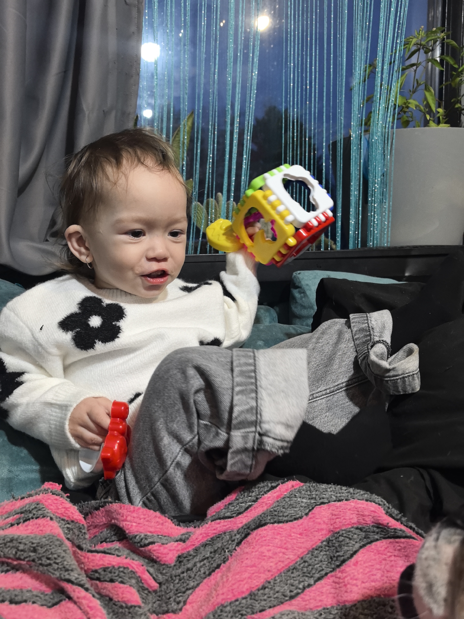 In the city of Noviy, Russia, a young child, approximately one to two years old, is seated indoors on a teal-colored upholstered surface, likely a sofa or armchair. The child has light brown hair and a visible small hoop earring in the right ear. They are wearing a white long-sleeved sweater adorned with multiple black floral patterns and grey denim jeans with cuffed ankles. A pink and grey striped blanket covers the child's lower body and lap. In the child's right hand, a brightly colored plastic toy, predominantly yellow with white, red, and green components, is held. In the left hand, a smaller, solid red, irregularly shaped plastic toy is present. The child's head is turned slightly to the right, and the mouth is partially open. Behind the child, a window is visible, partially covered by light blue string curtains. A grey fabric curtain hangs to the left of the window. Outside the window, out-of-focus lights suggest an evening or nighttime scene. A potted green plant is visible in the background to the right of the window. A dark-colored cushion is situated near the child's right side.